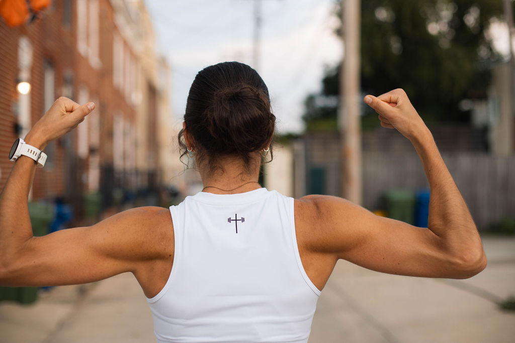 A girl wearing a white Christian Workout top that has a cross with a barbell flexing