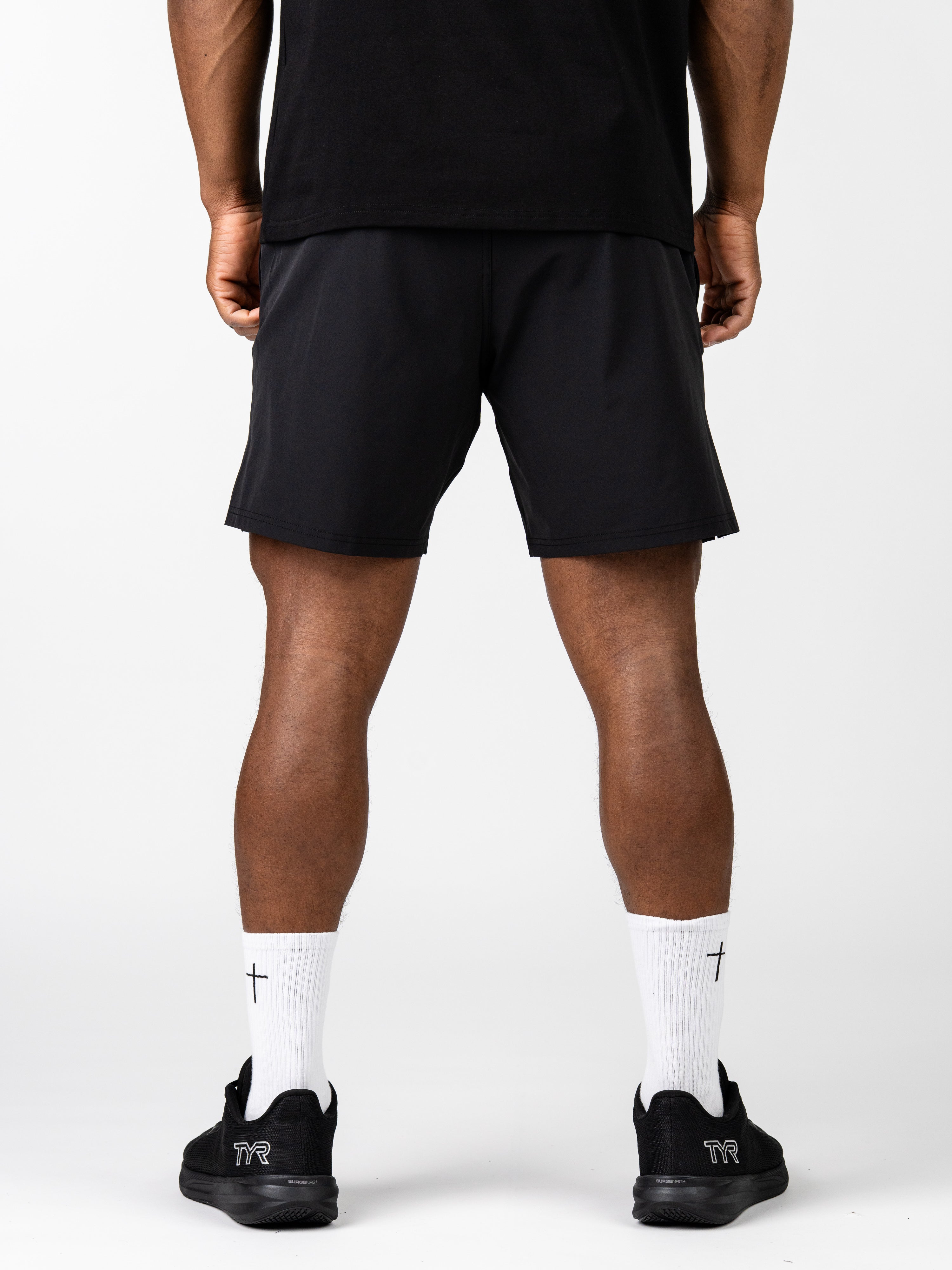 Rear view of a person wearing black Essential Training Shorts paired with white crew socks featuring a small cross design and black athletic shoes, showcasing lightweight, breathable Christian gym clothing.