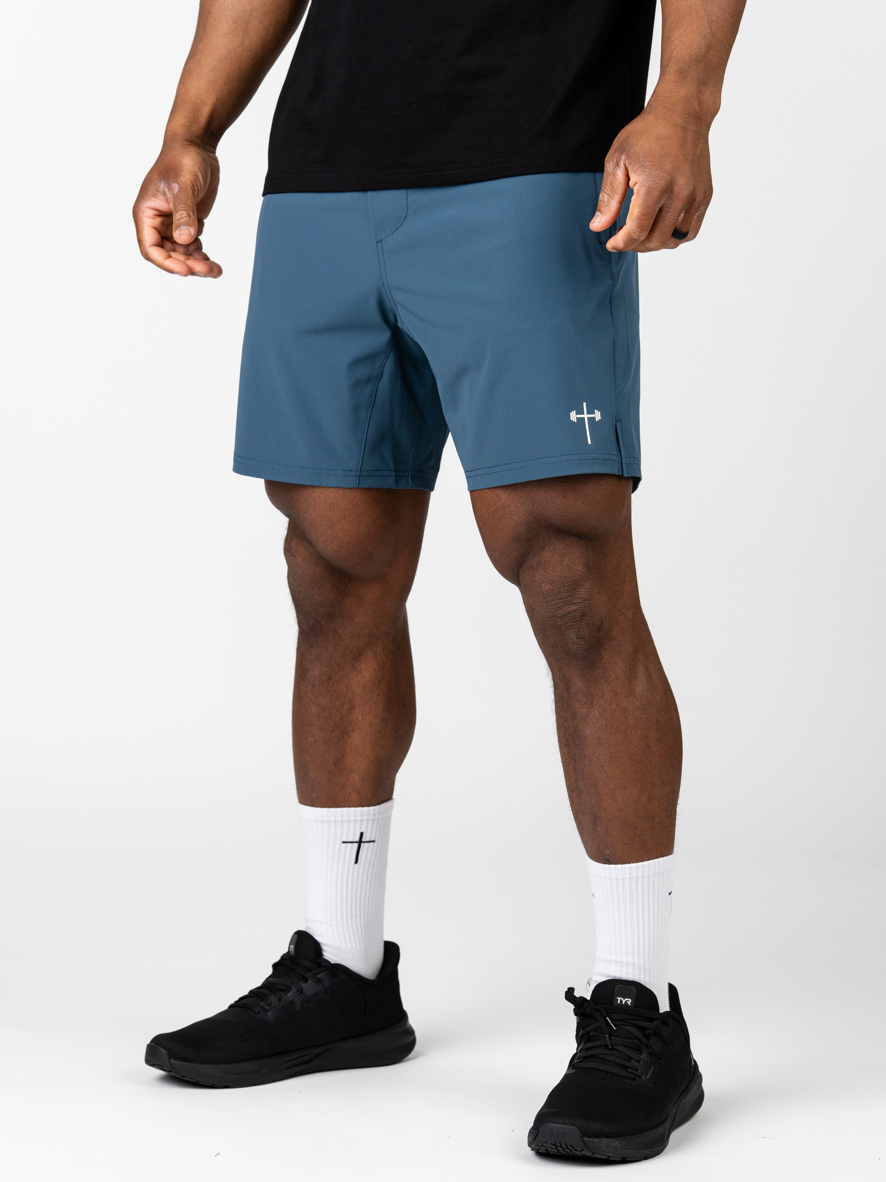 Male model wearing blue Essential Training Shorts with a small white cross and barbell logo on the leg, paired with white socks featuring a cross and black athletic shoes.