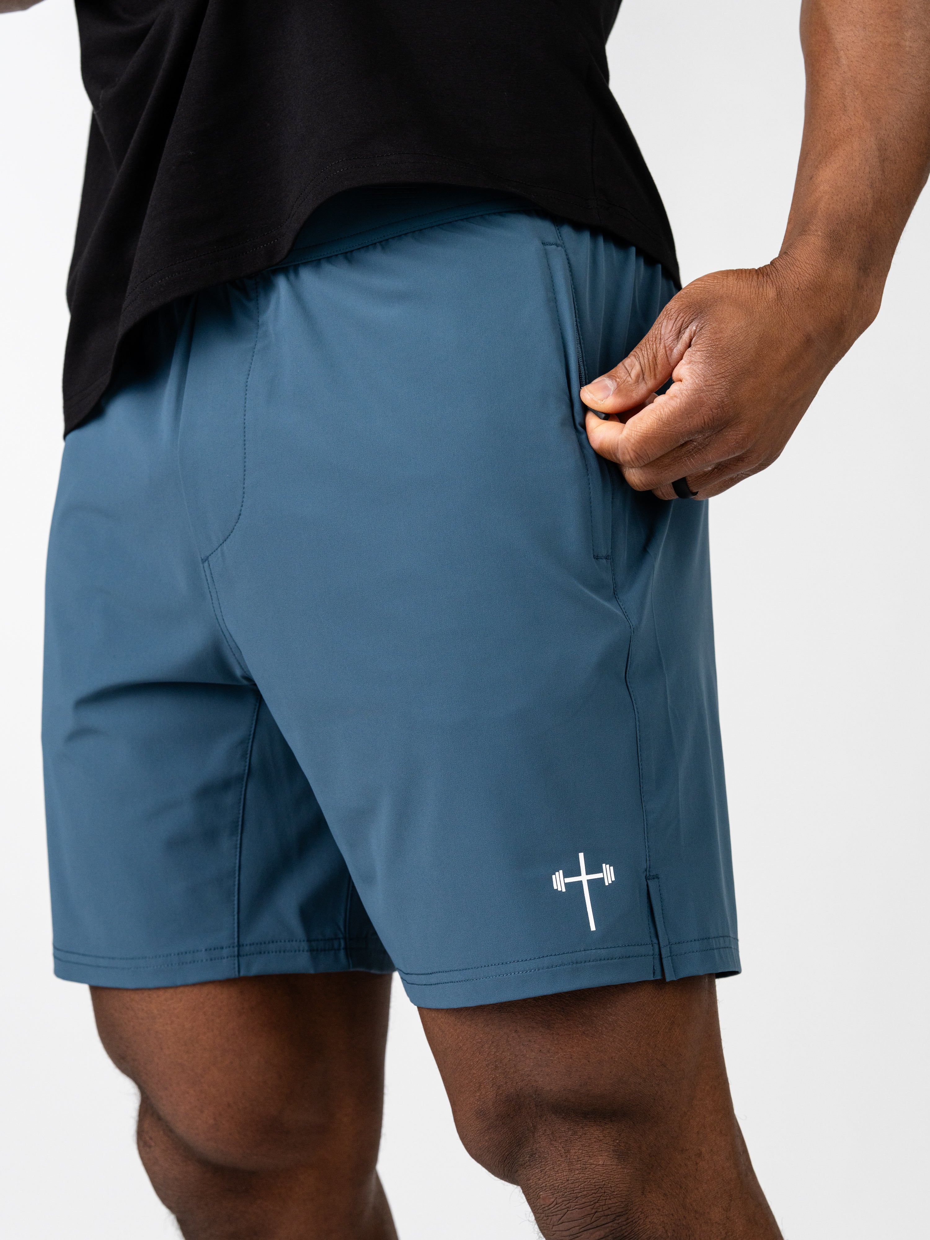 Close-up of a person wearing blue Essential Training Shorts with a small white cross and barbell logo near the hem, showing the lightweight fabric and side pocket.