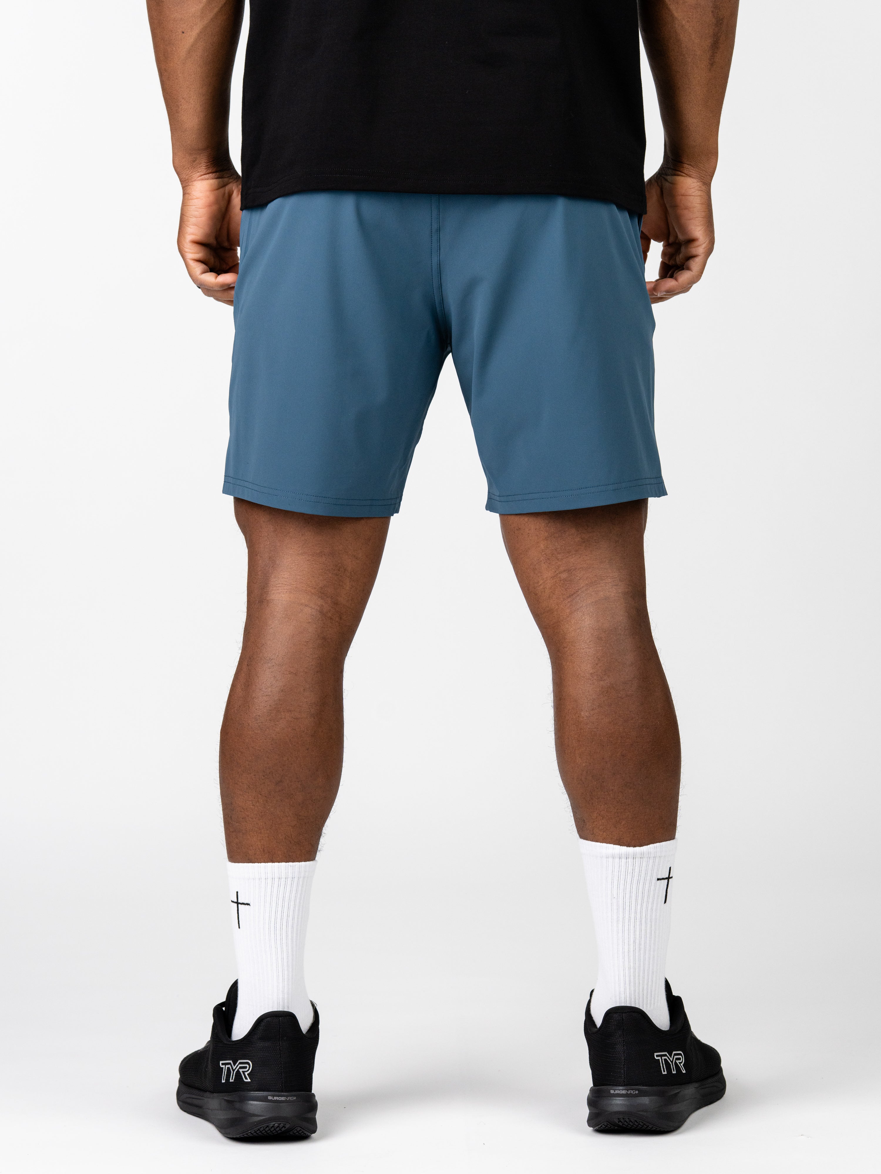 Back view of a man wearing blue Essential Training Shorts paired with white socks featuring a subtle cross design and black athletic shoes, showcasing lightweight, breathable Christian workout clothes.