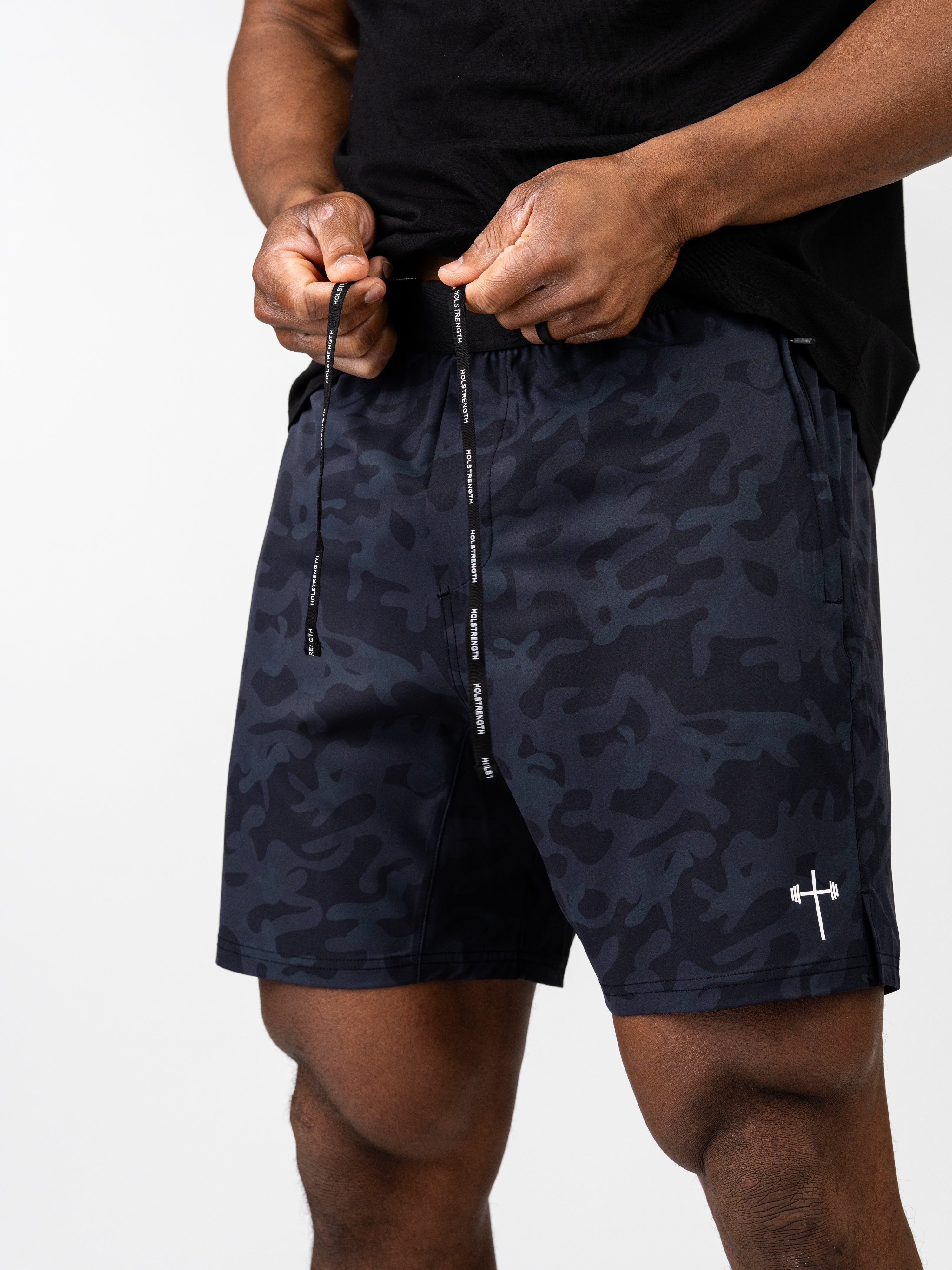 Man adjusting black camo Essential Training Shorts with drawstring and discreet logo on left leg, designed for comfort and movement during workouts.