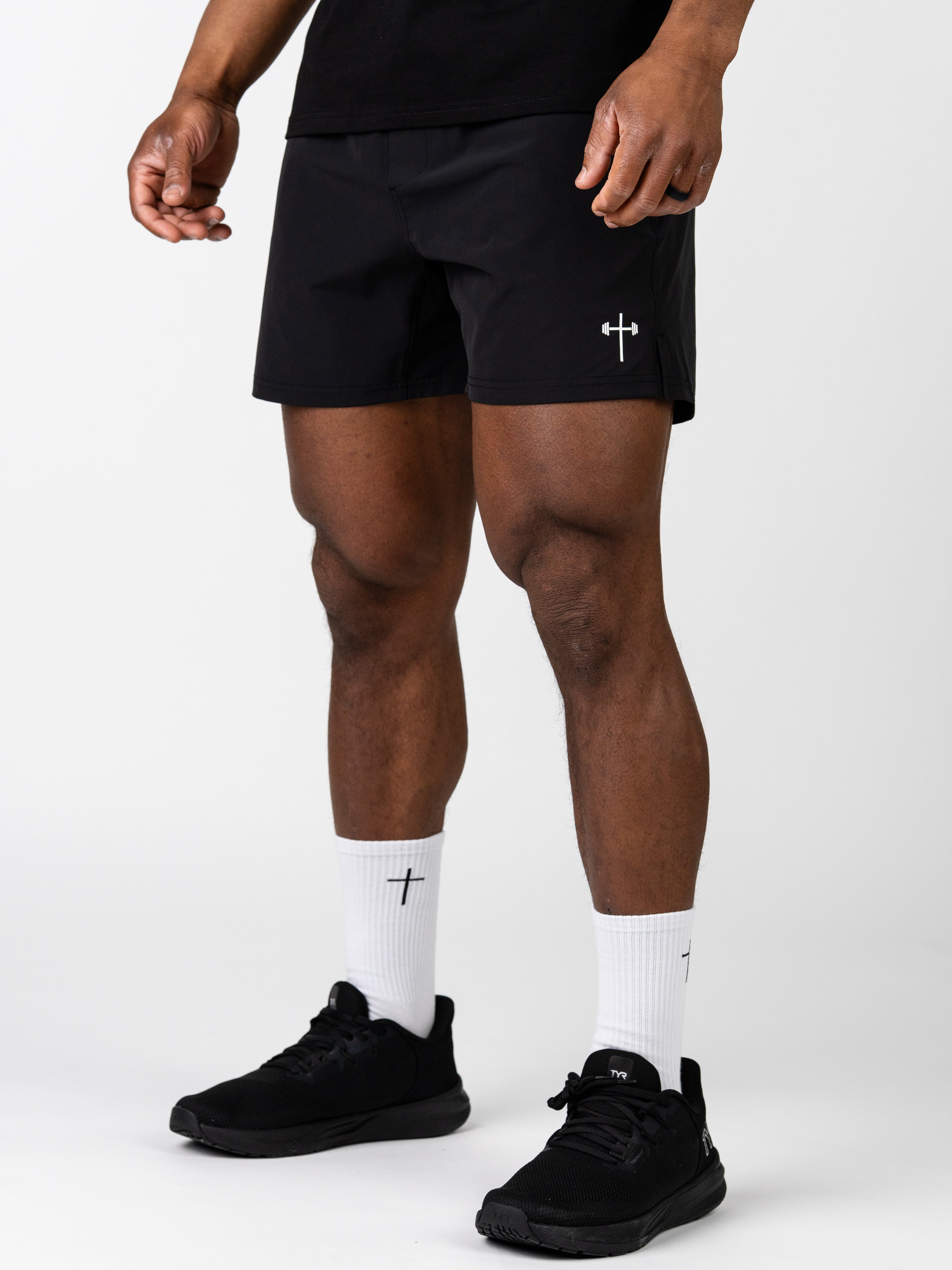 Front view of a person wearing black Essential Training Shorts with a small white cross and barbell logo on the left leg, paired with white cross-logo socks and black athletic shoes.