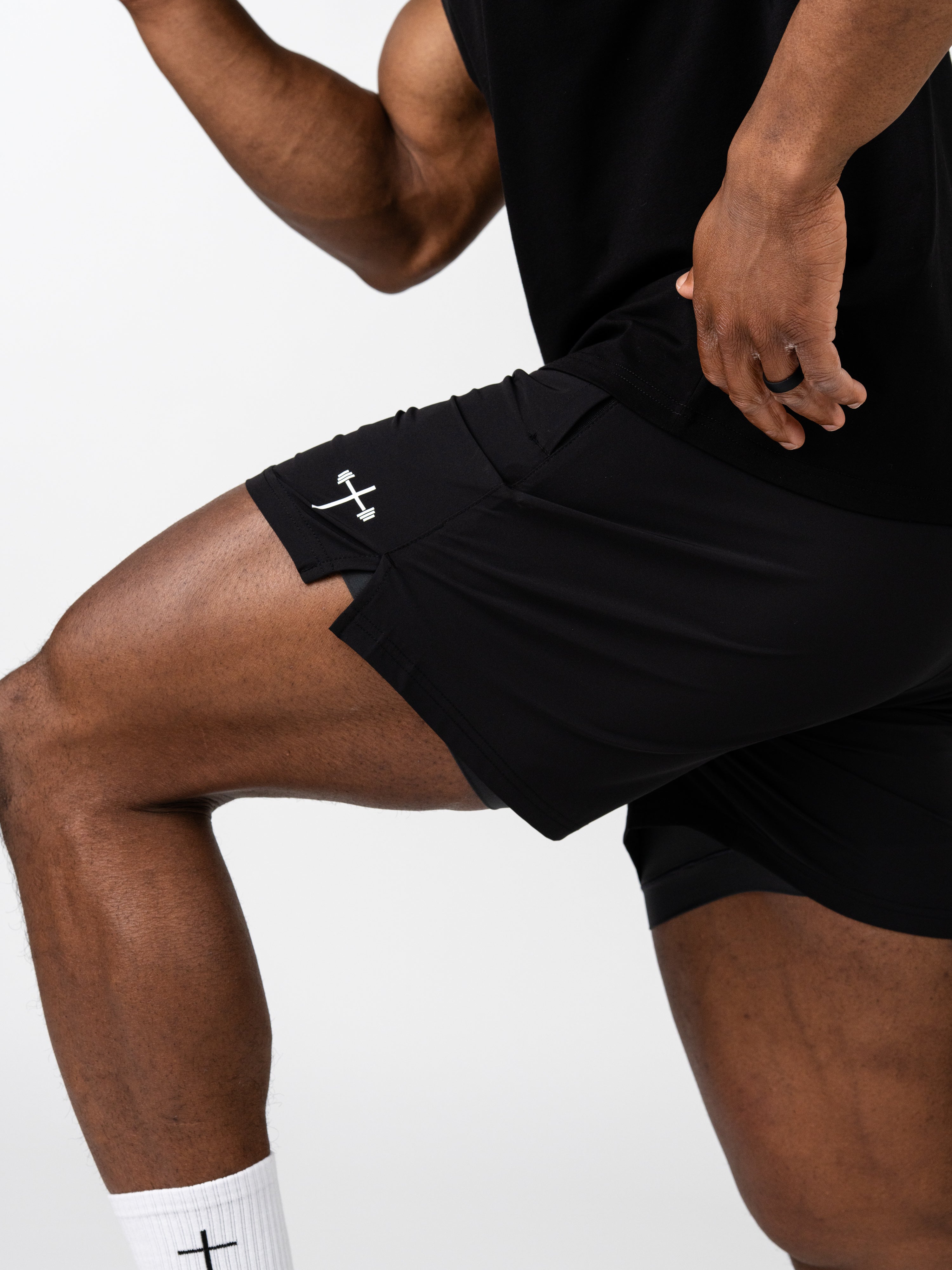 Athletic person wearing black Essential Training Shorts featuring a small white cross and barbell symbol on the side, paired with white socks with cross design, in a dynamic workout pose.