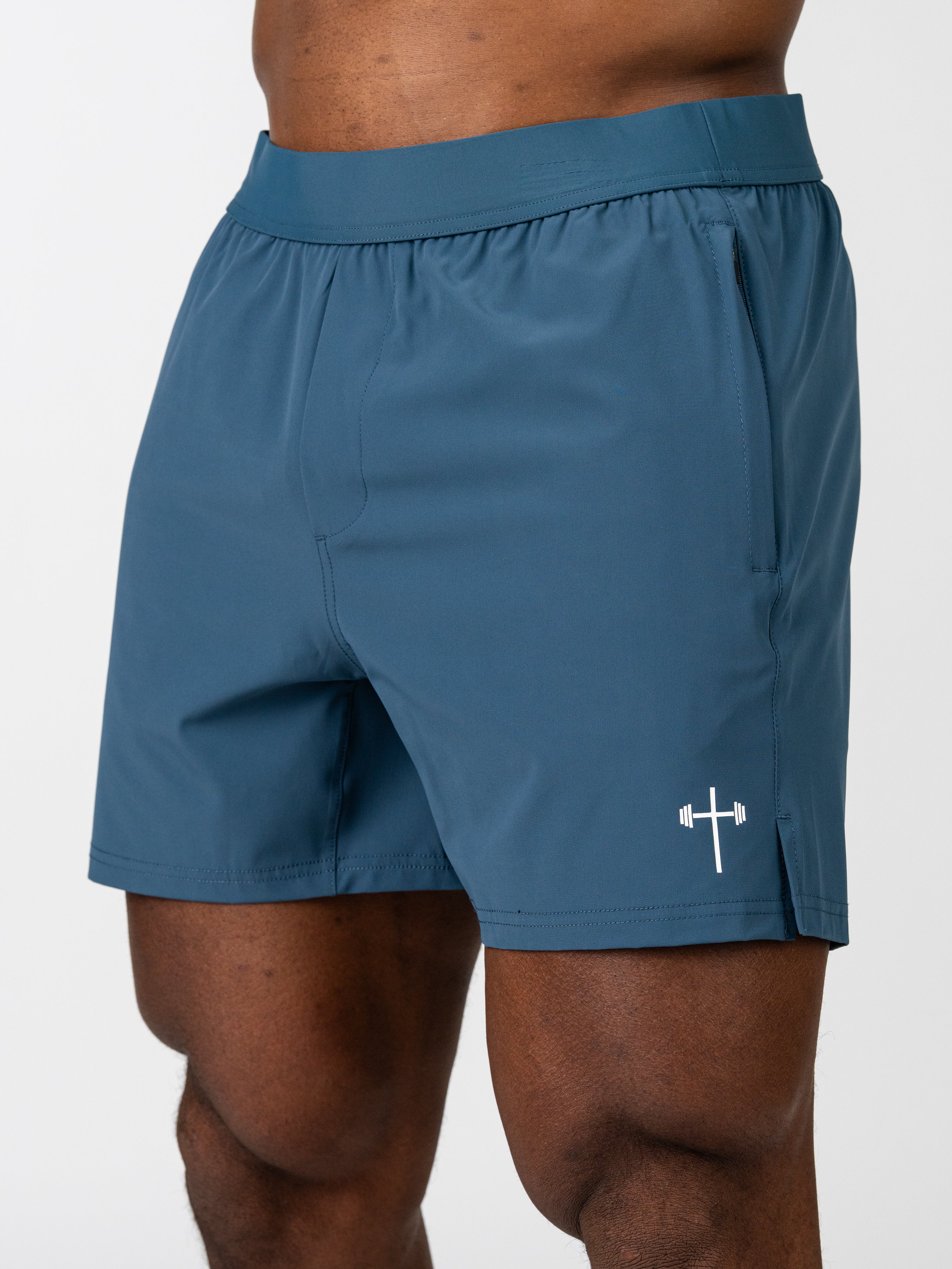 Male model wearing blue Essential Training Shorts featuring a small white cross with a barbell design near the hem, designed for comfortable movement during workouts.