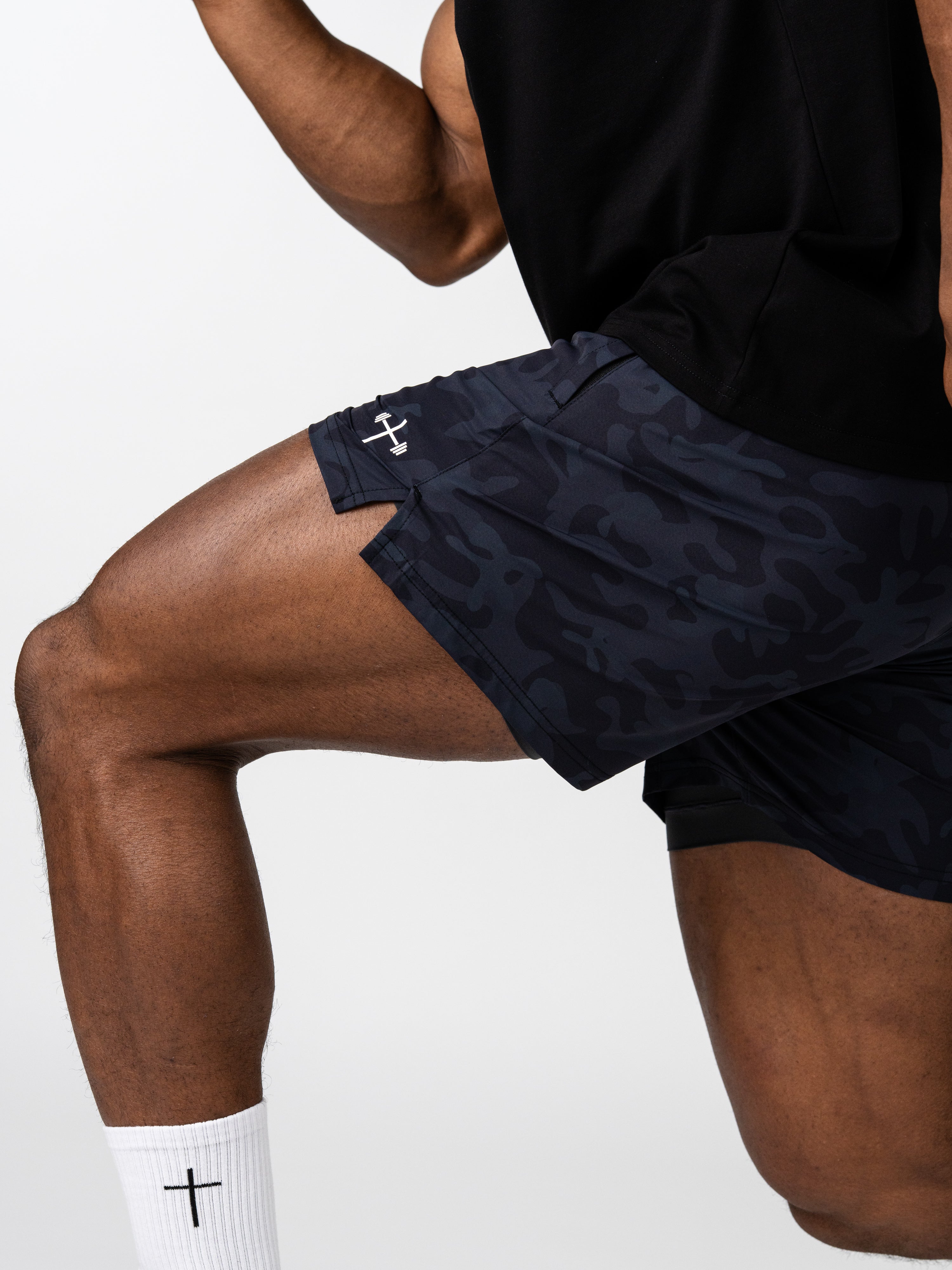 Athlete wearing Essential Training Shorts with dark camouflage pattern and small white Christian cross emblem, paired with white socks featuring a cross design.