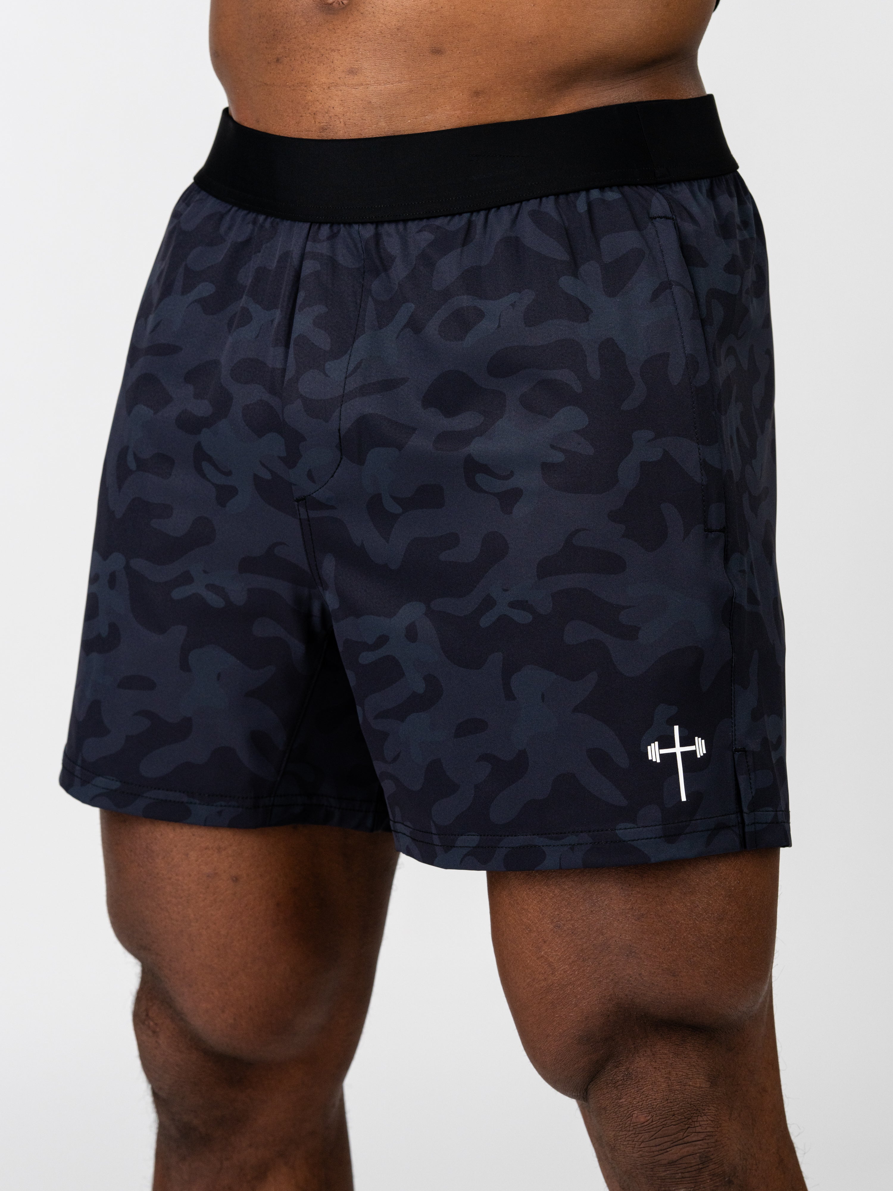 Close-up of a person wearing Essential Training Shorts featuring a dark camo pattern with a black waistband and a small white cross and barbell logo on the lower left leg.