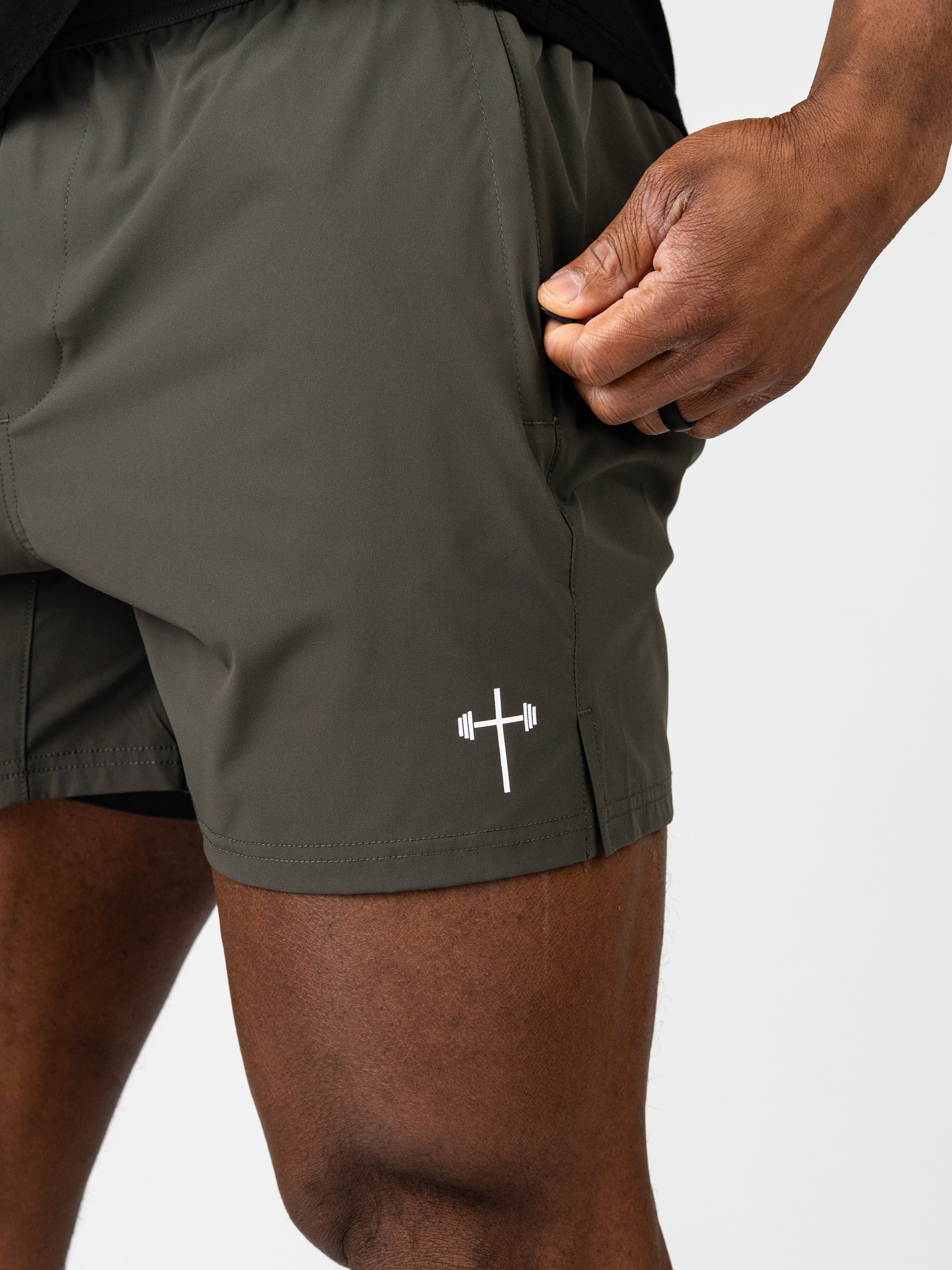 Close-up of a person wearing olive green Essential Training Shorts with a white Christian cross and barbell logo near the hem, featuring a convenient side pocket.