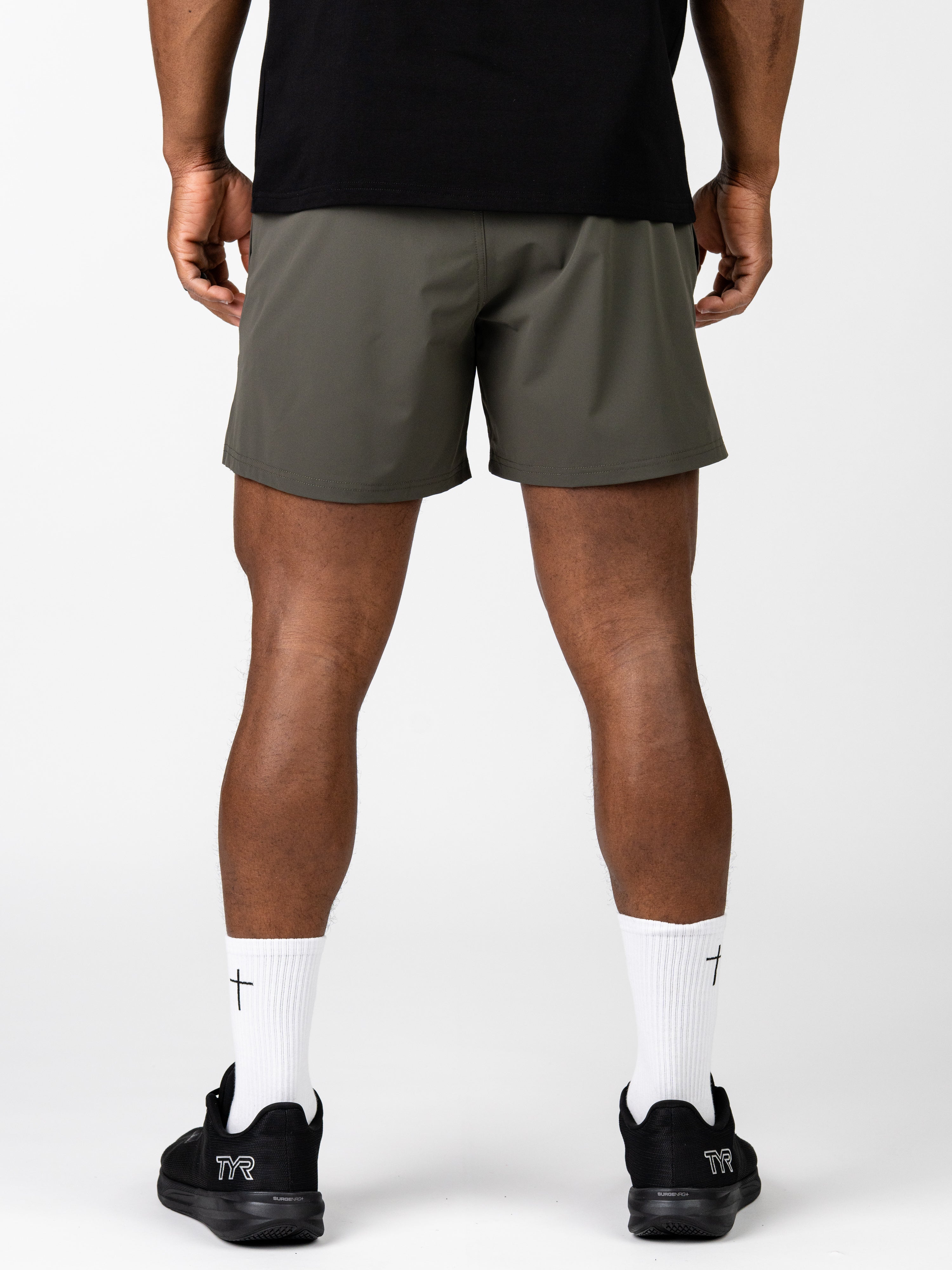 Man wearing olive green Essential Training Shorts standing with back facing camera, paired with white socks featuring small crosses and black athletic shoes.