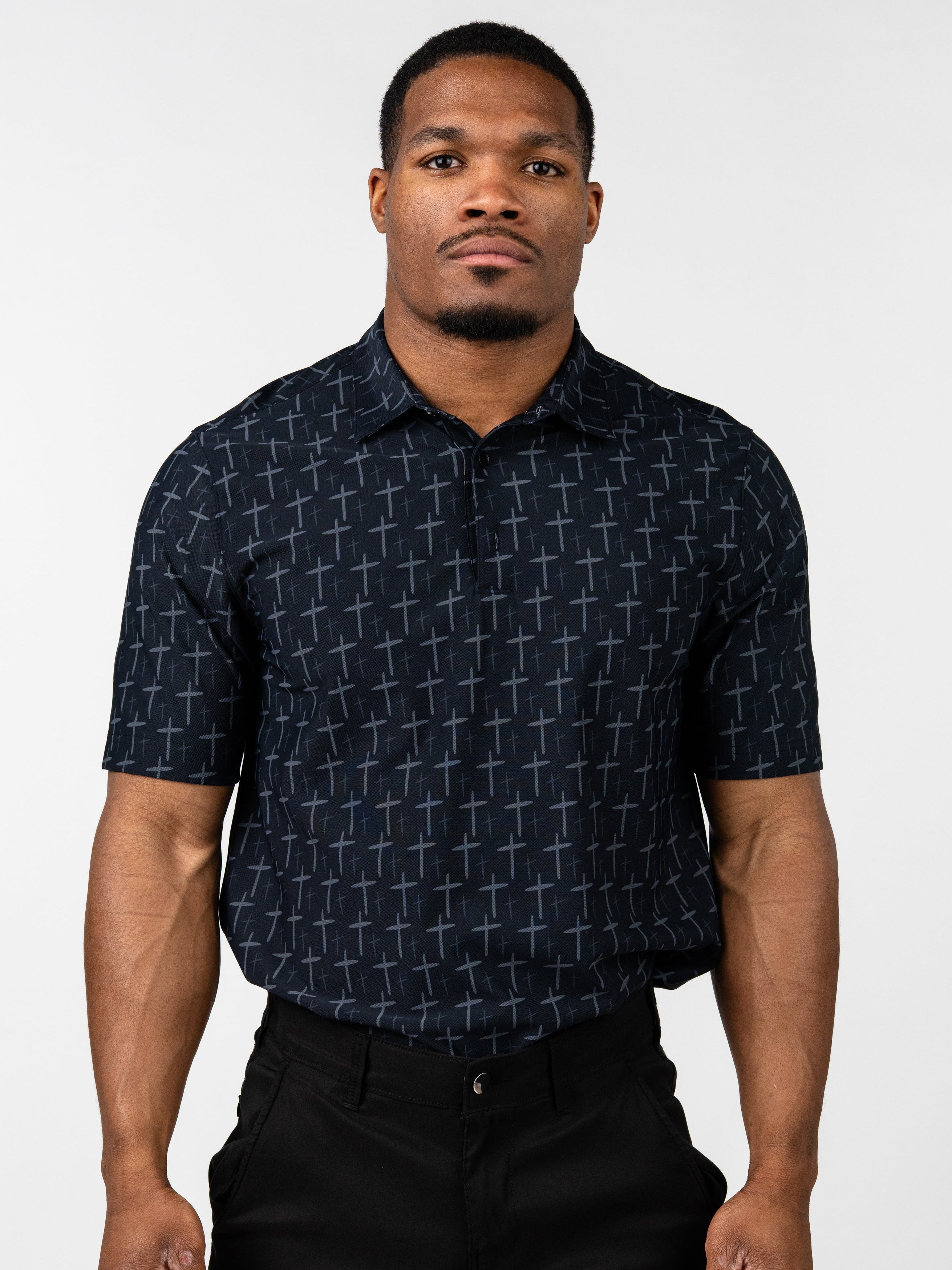 Man wearing a black Cross Premium Polo shirt patterned with subtle gray crosses, paired with black pants, standing against a plain white background.