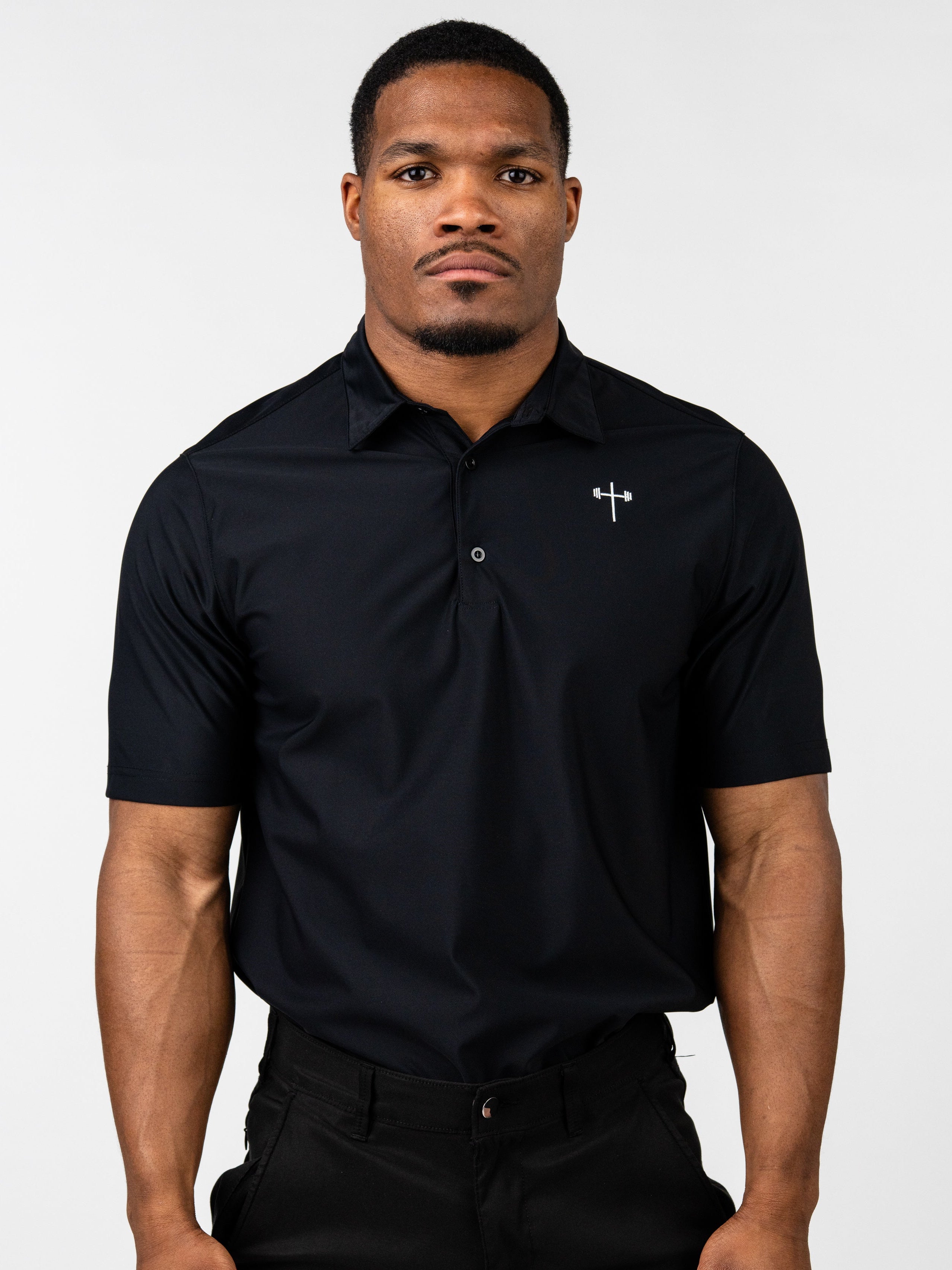 Male model wearing a black Core Athletic Polo with a subtle Christian cross and barbell logo on the chest, styled with black pants against a plain background.