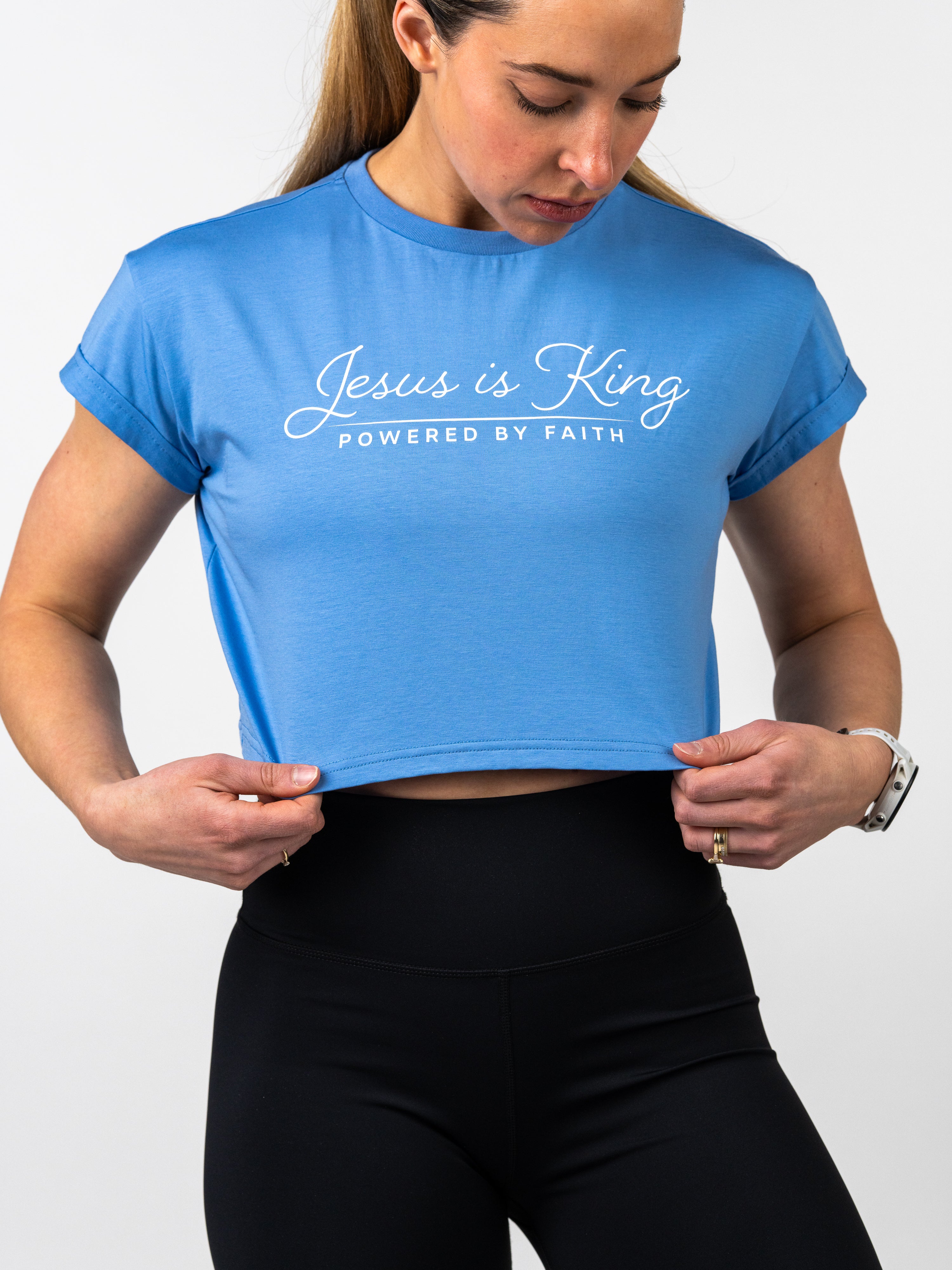 Woman wearing a blue Jesus is King Essential Crop Tee with white lettering, paired with black high-waisted leggings, showcasing a sporty and faith-based outfit.