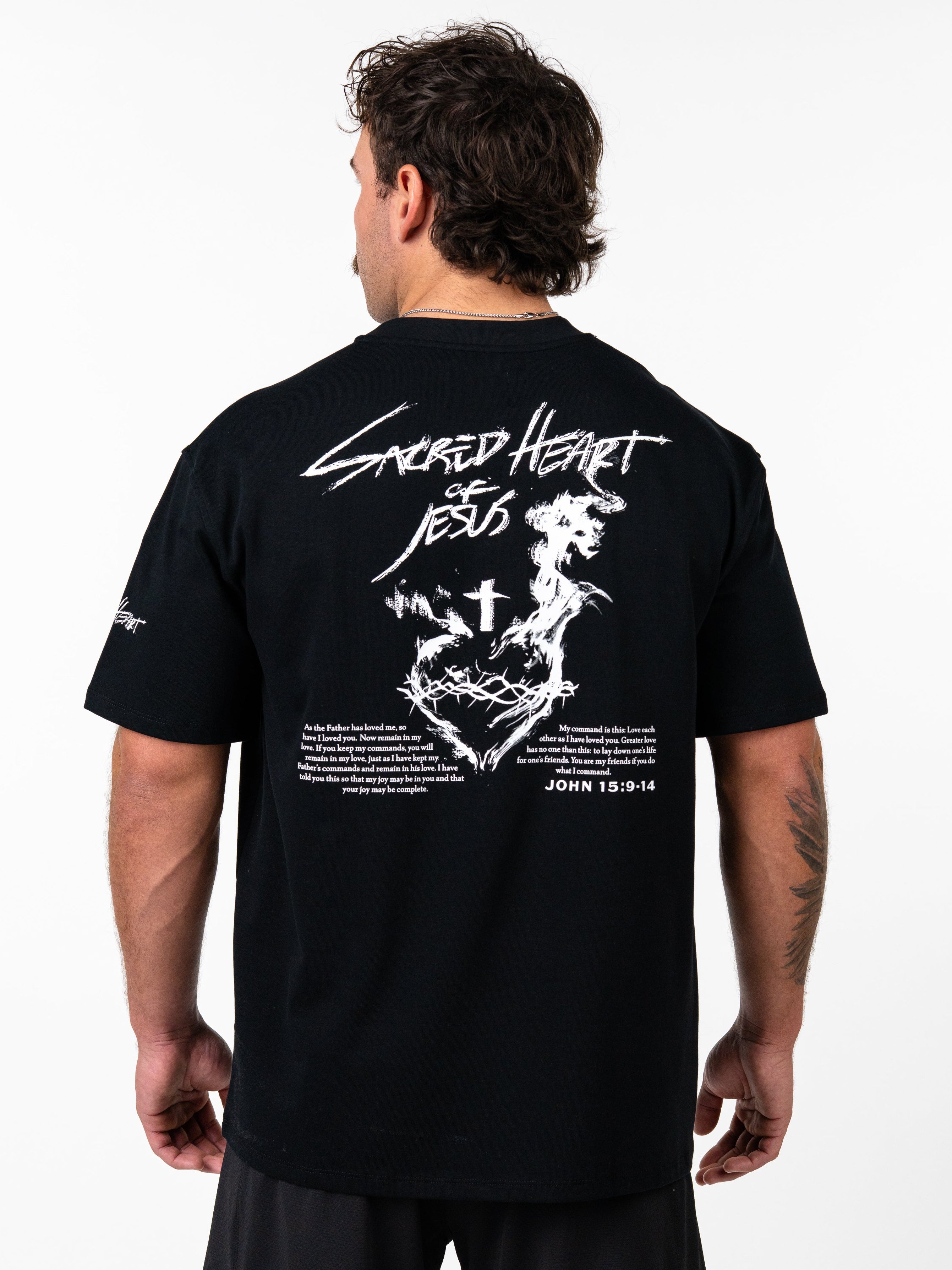 Man wearing black Sacred Heart Premium Oversized Tee with white graphic of a crowned heart, cross, barbell design, and biblical text on the back, styled as Christian fitness apparel.