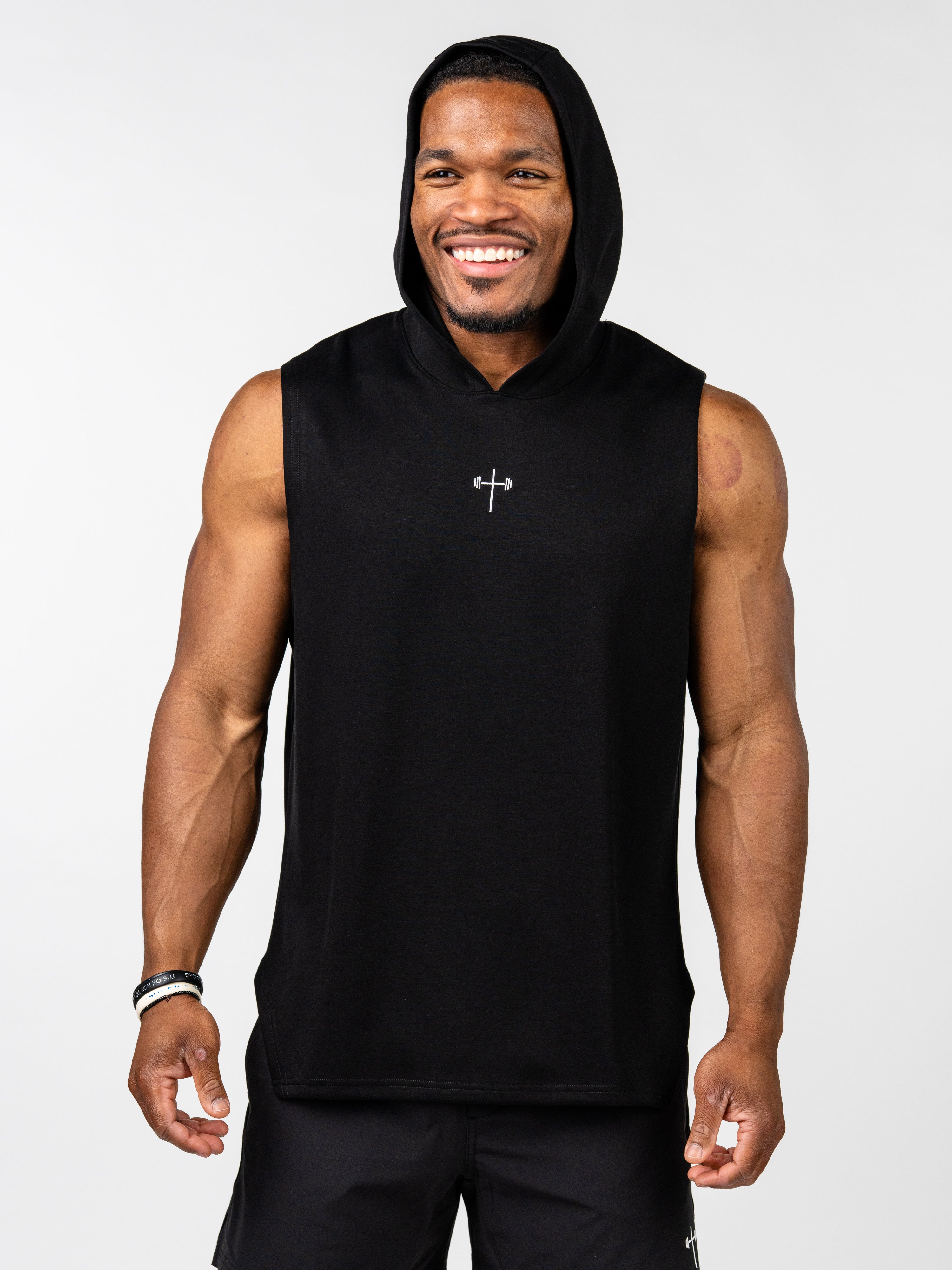 Male athlete wearing a black Essential Sleeveless Hoodie with a subtle silver cross and barbell design on the chest, smiling while standing against a plain background.