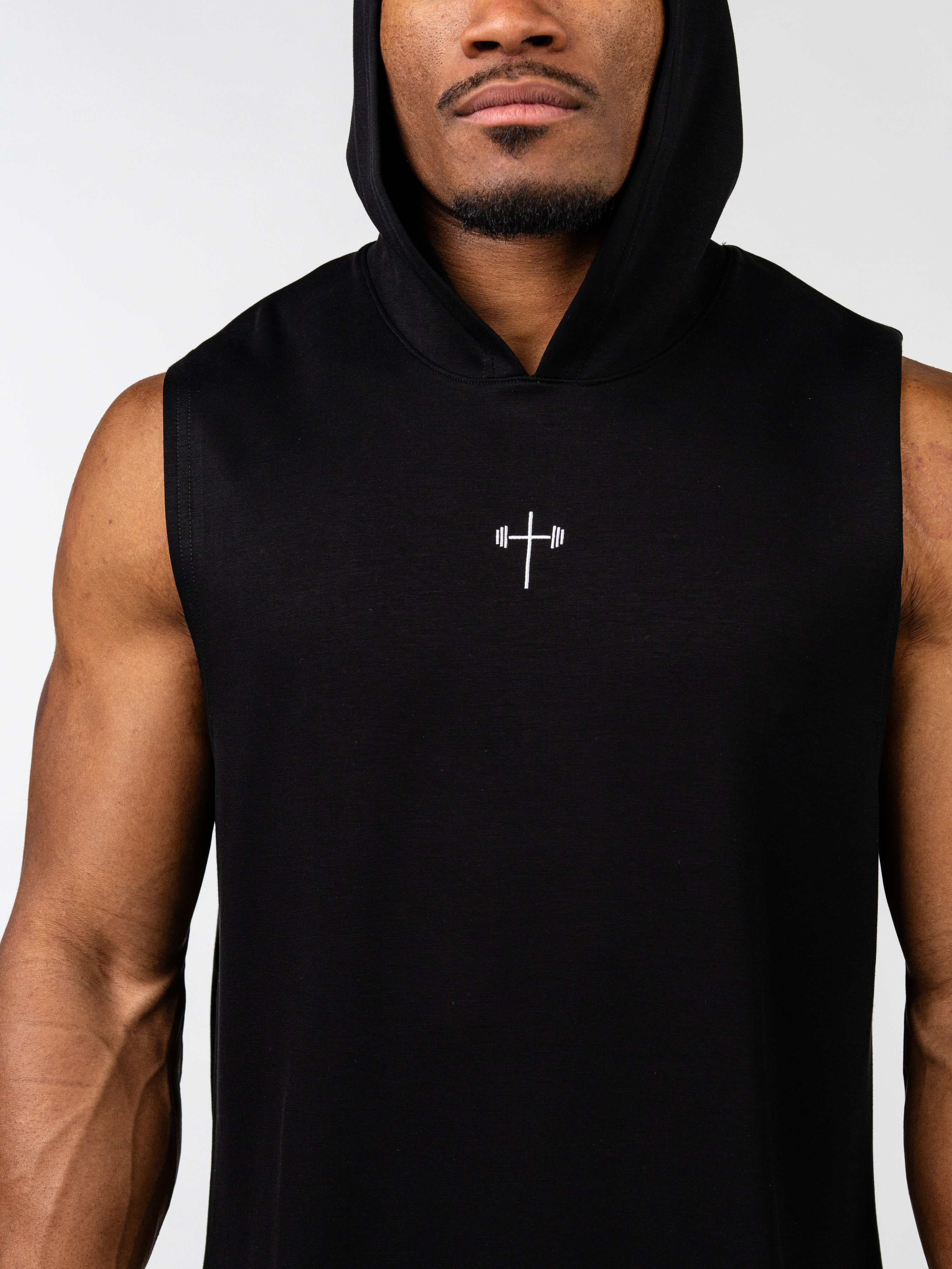 Close-up of a man wearing a black Essential Sleeveless Hoodie featuring a small white cross with a barbell design on the chest, representing Christian fitness apparel.
