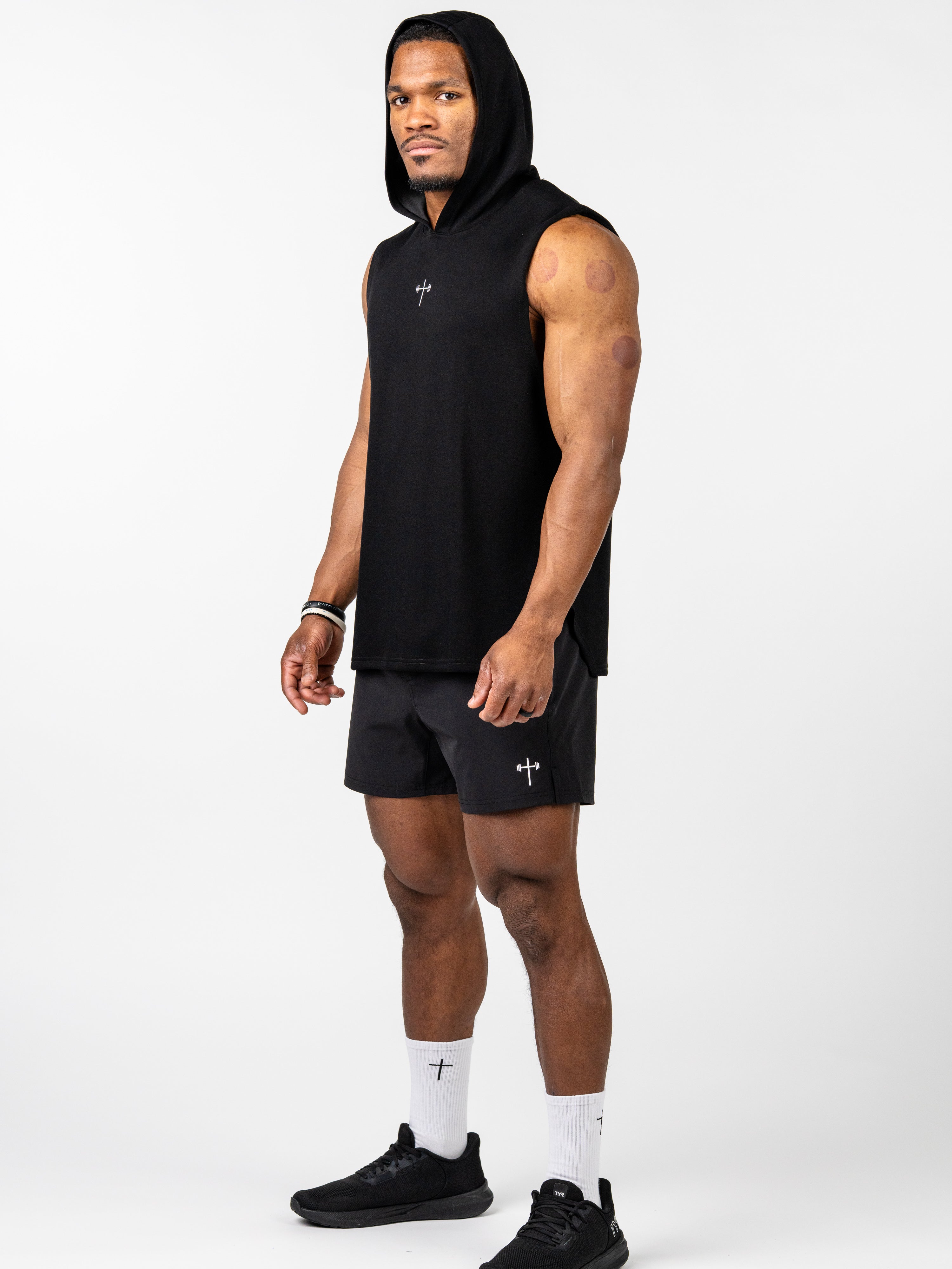 Athletic male model wearing a black Essential Sleeveless Hoodie with a small cross and barbell logo, paired with matching shorts and white socks featuring a cross design.