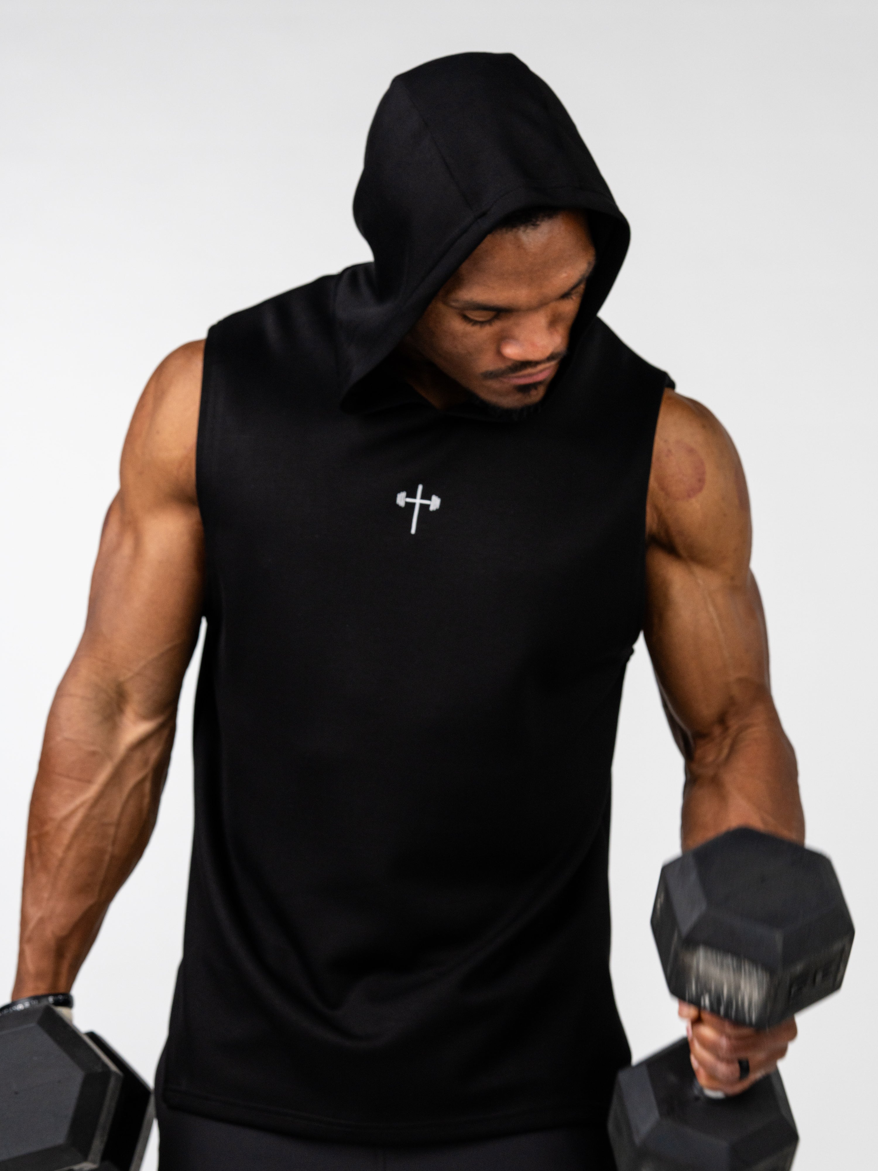 Man wearing a black Essential Sleeveless Hoodie with a small cross and barbell logo on the chest, lifting dumbbells in a gym setting.