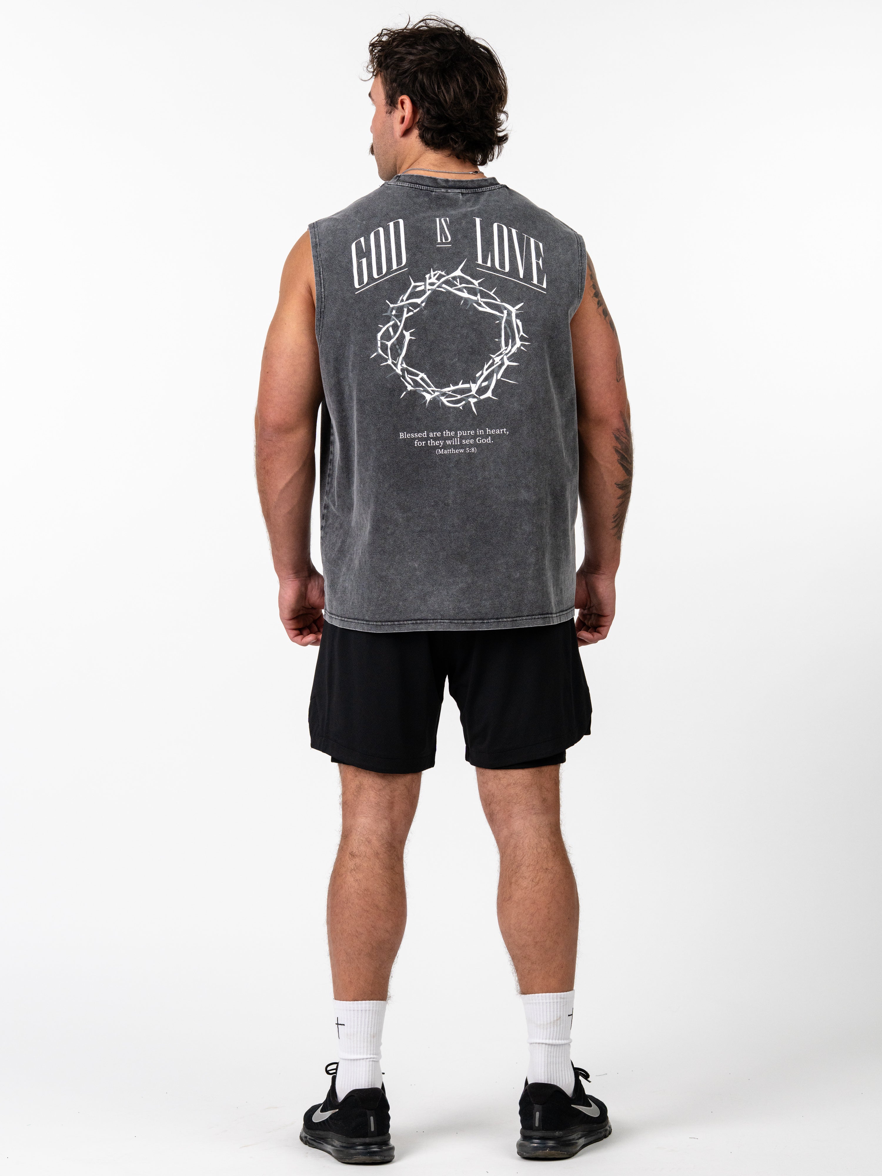 Man wearing a sleeveless gray God is Love Vintage Premium Tank with a crown of thorns graphic and scripture text on the back, paired with black shorts and athletic shoes.