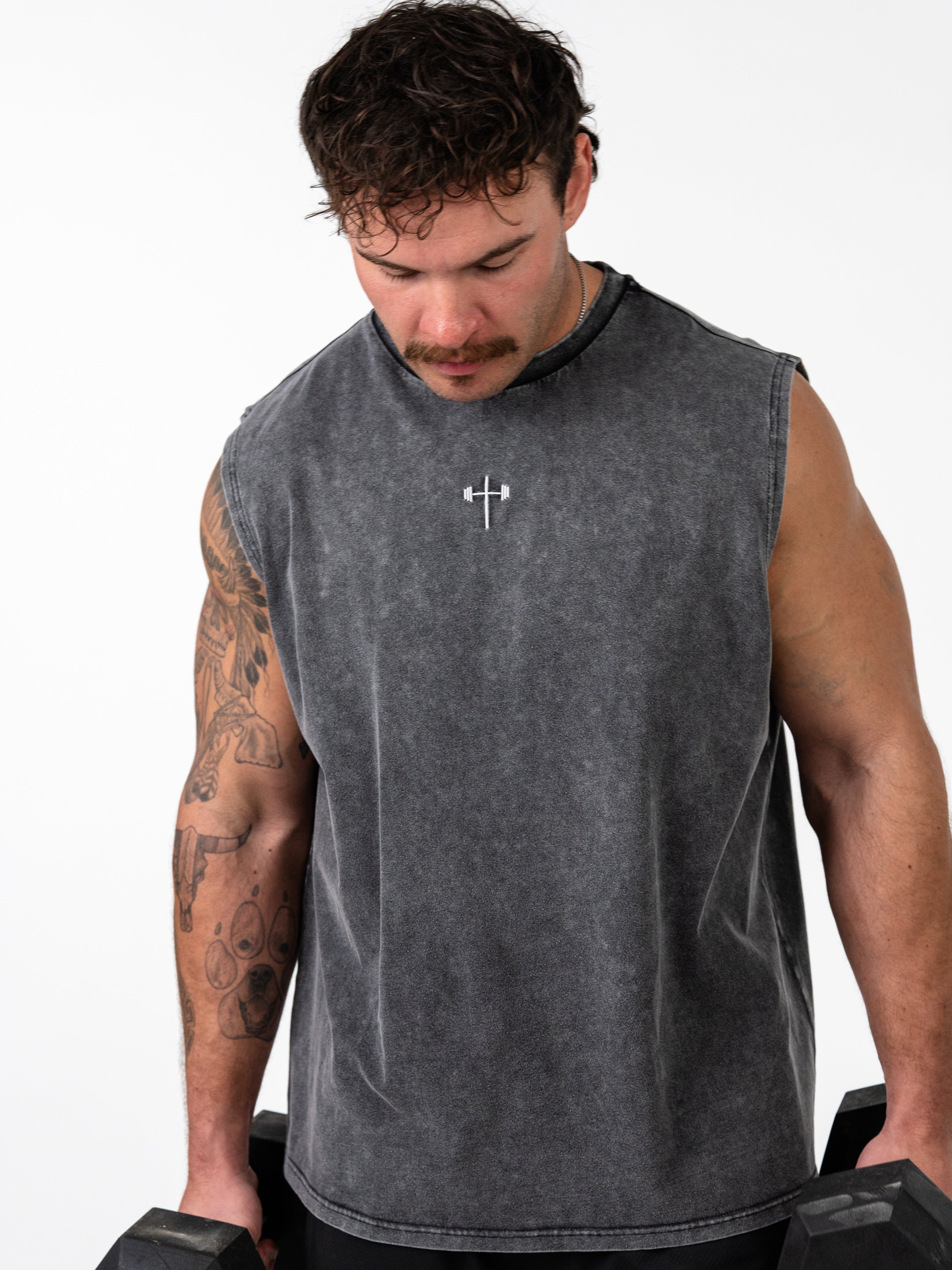 Man wearing a dark gray sleeveless gym tank with a small white barbell cross symbol on the chest, lifting dumbbells, wearing the Servant of God Vintage Premium Tank.