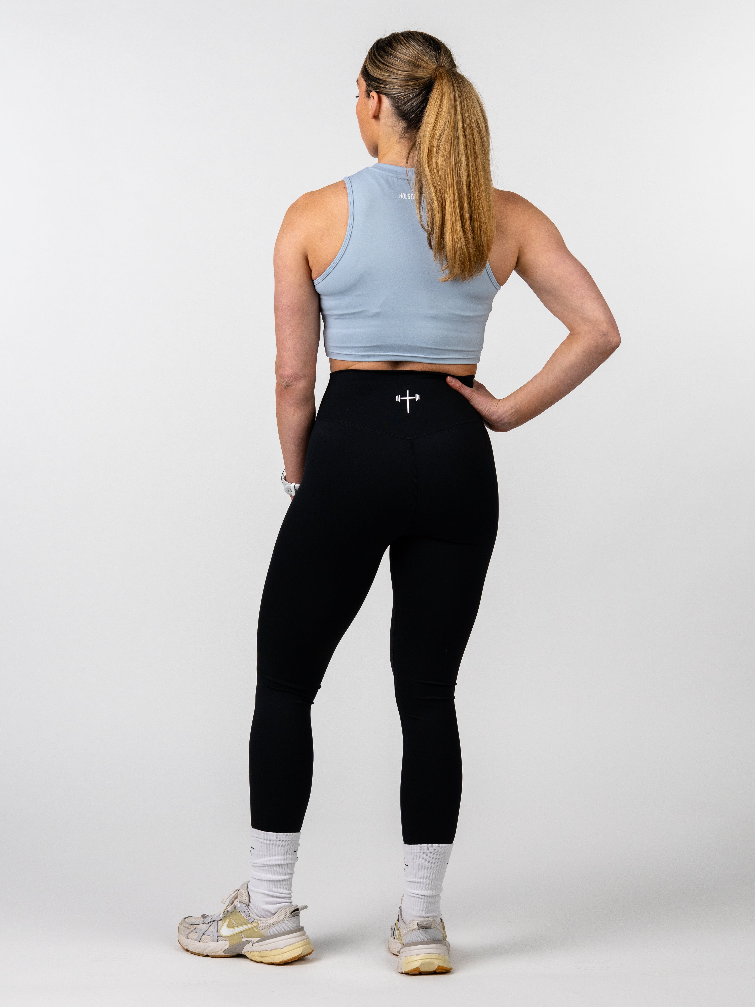 Woman wearing a Vital Cross Combat Top shown from behind, paired with black leggings featuring a small white cross and barbell design at the waistband, demonstrating Christian fitness apparel style.