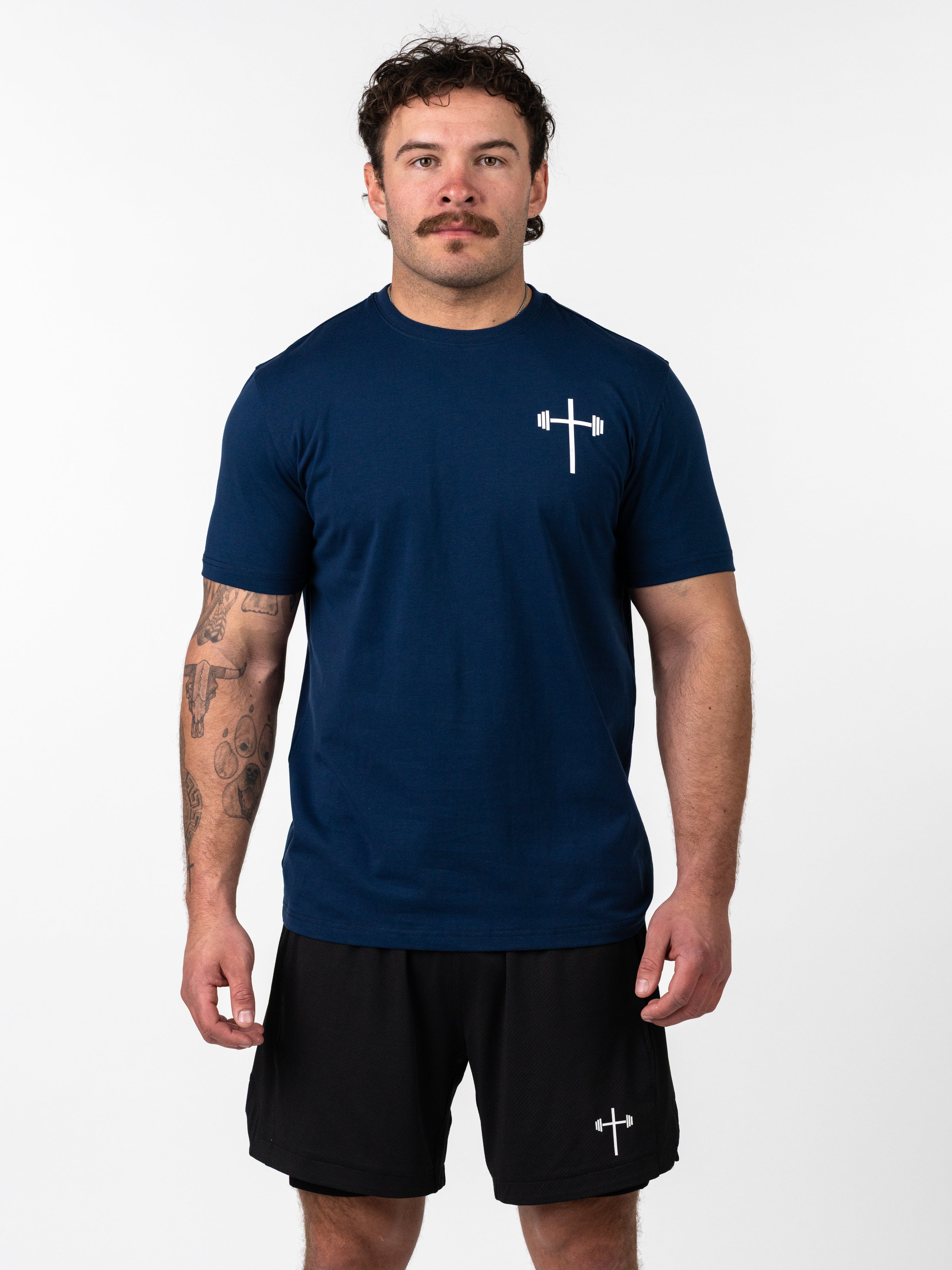 Man wearing a navy blue Kingdom Performance Tee with a small white barbell cross logo on the chest, paired with black shorts featuring a matching logo, showcasing Christian fitness apparel.