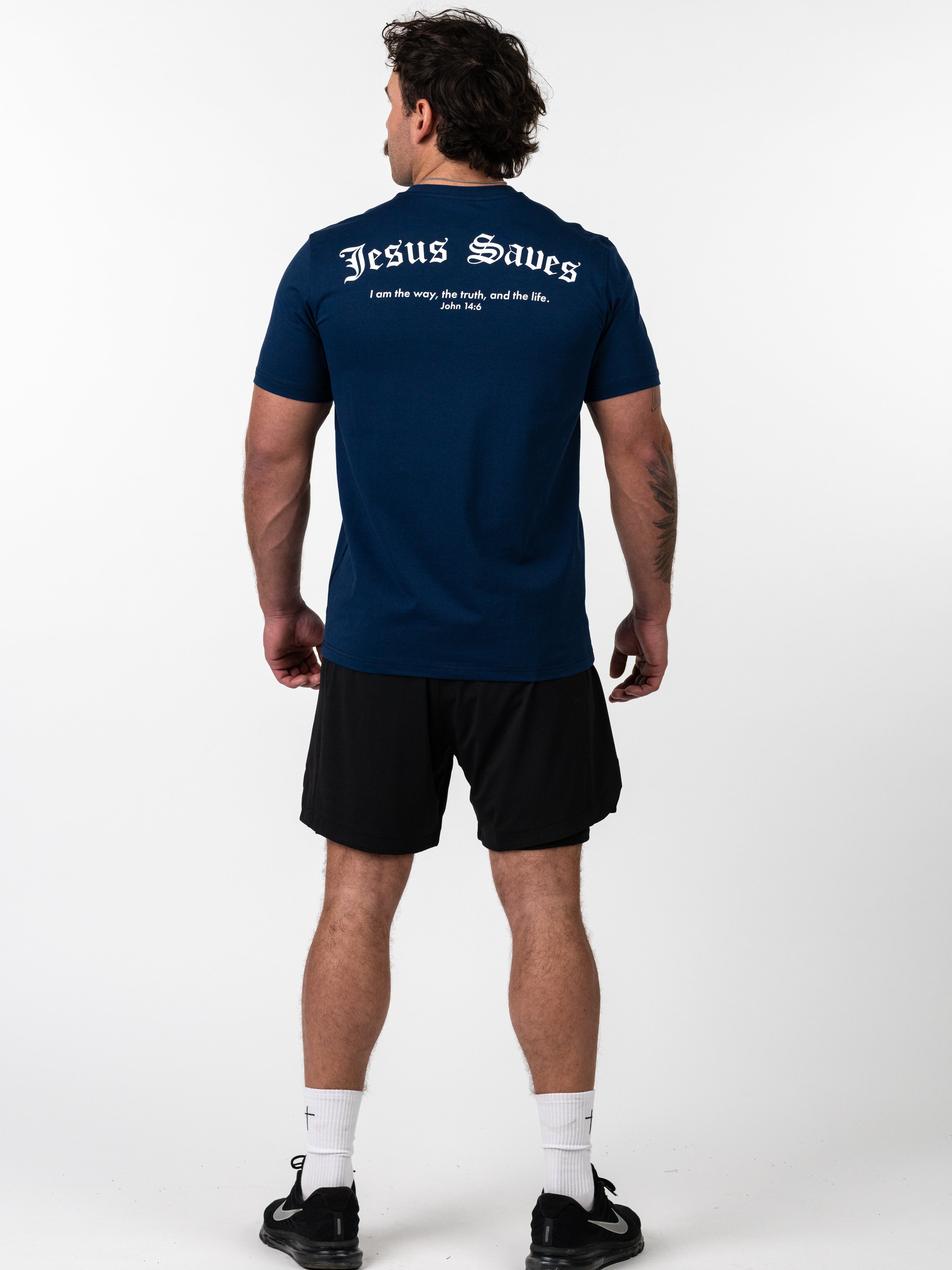 Man wearing a navy blue Kingdom Performance Tee with "Jesus Saves" and a Bible verse printed on the back, paired with black shorts and athletic shoes.