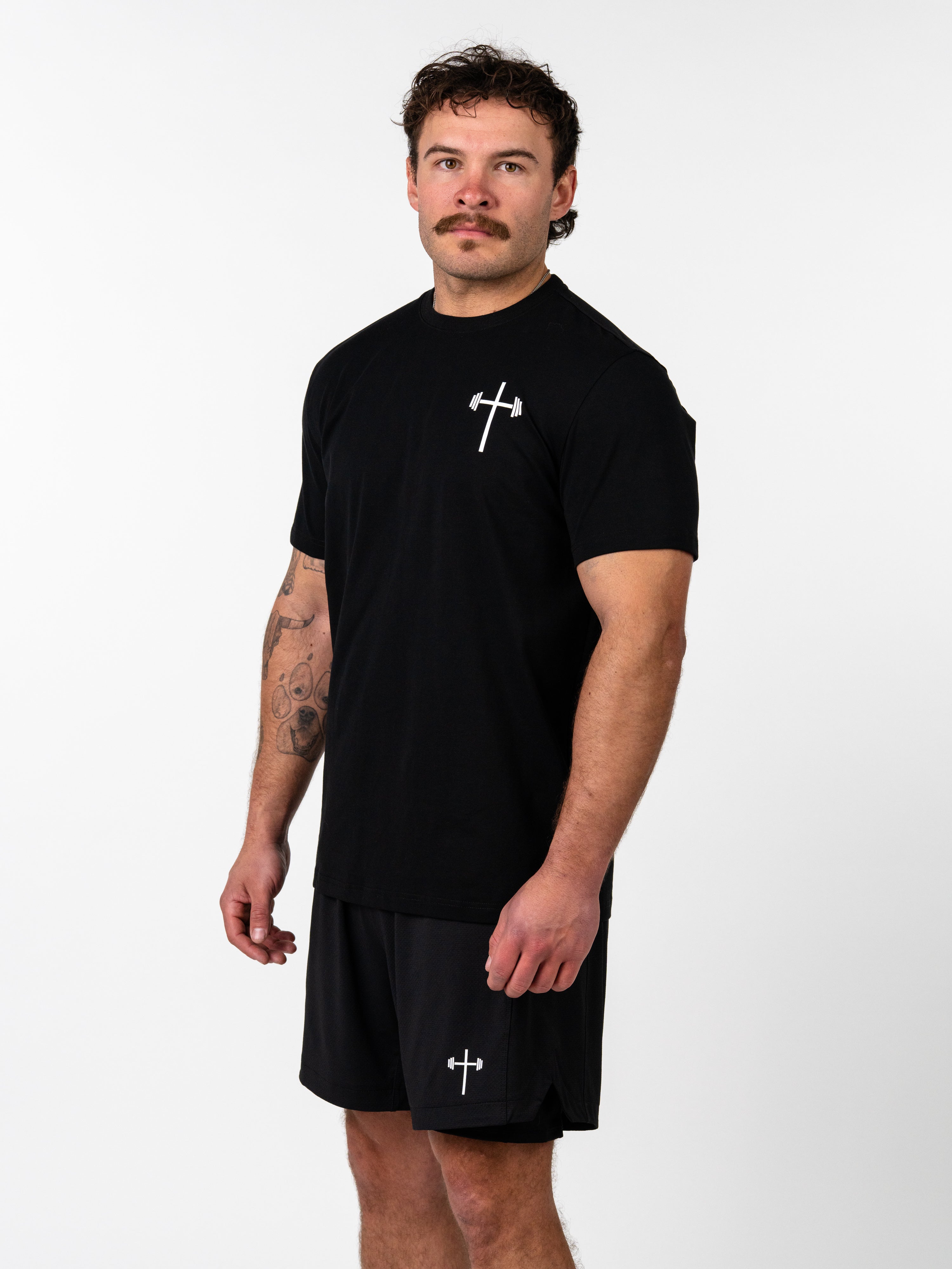 Male model wearing a black Kingdom Performance Tee featuring a small white barbell and cross logo on the chest, paired with matching black shorts, showcasing Christian gym clothing.
