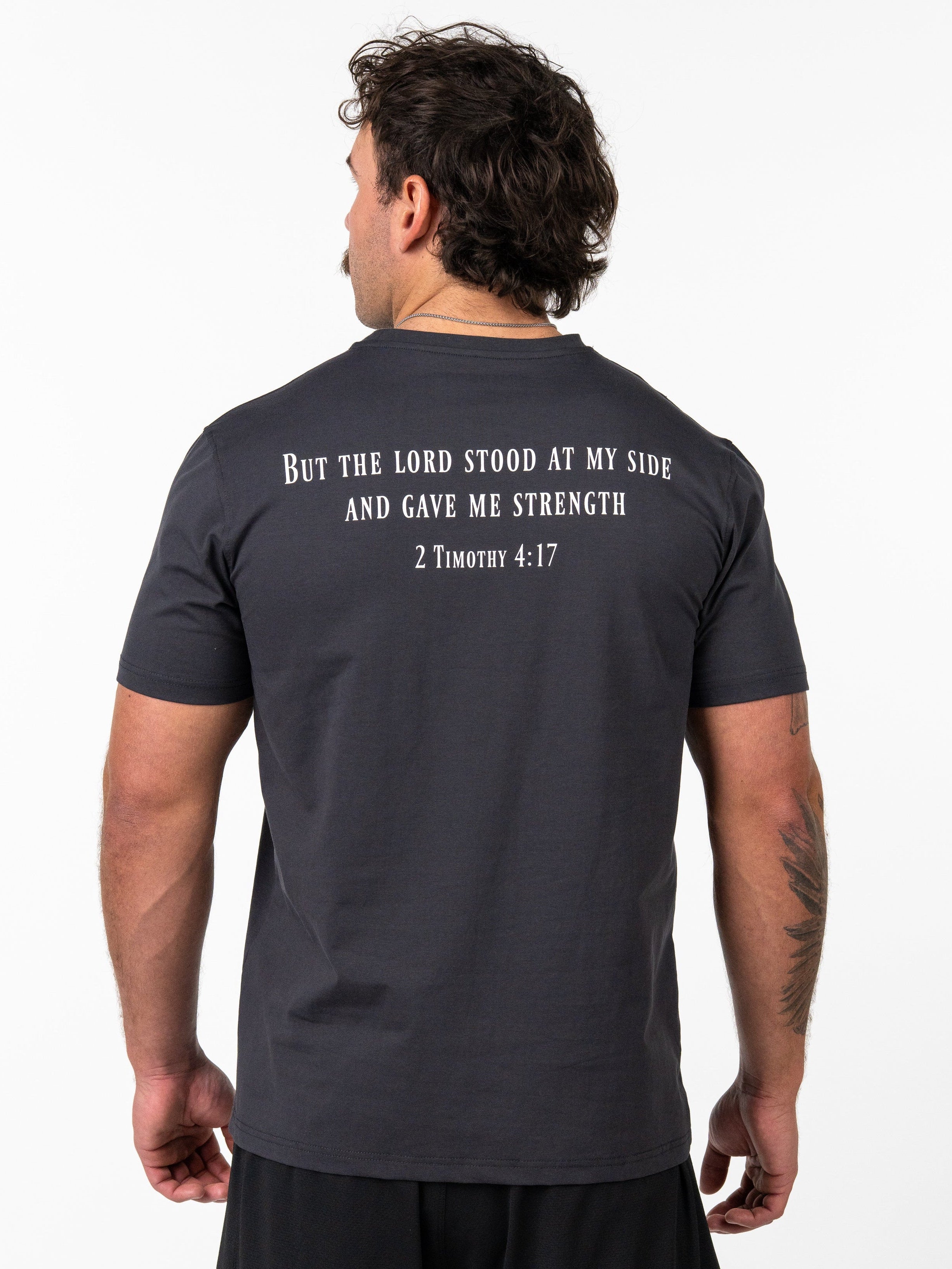 Man wearing a black Sovereign Performance Tee with a biblical quote from 2 Timothy 4:17 printed on the back, paired with black athletic shorts in a studio setting.