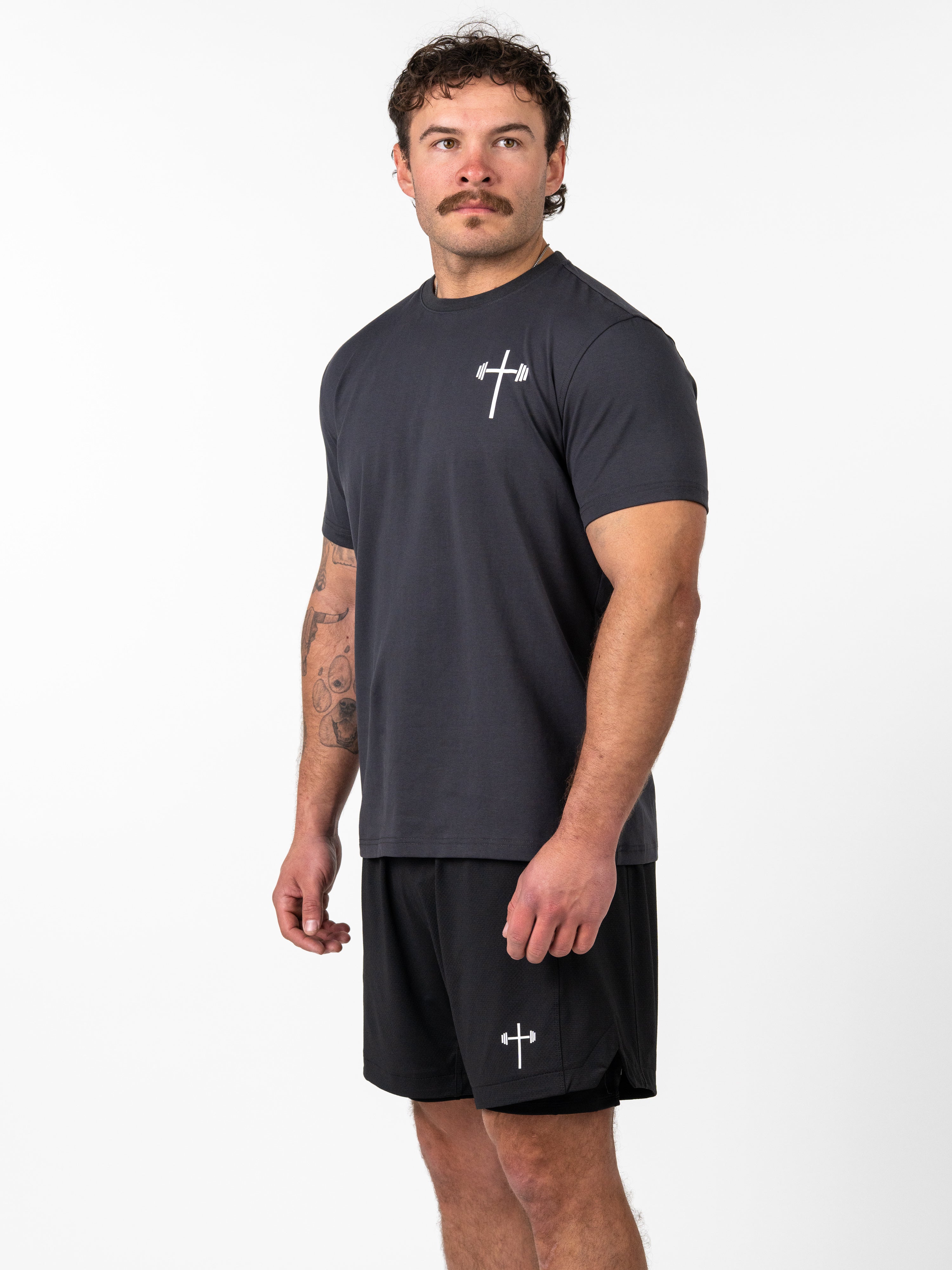 Male model wearing a black Kingdom Performance Tee with a white cross and barbell graphic on the chest, paired with matching black shorts, standing against a white background.
