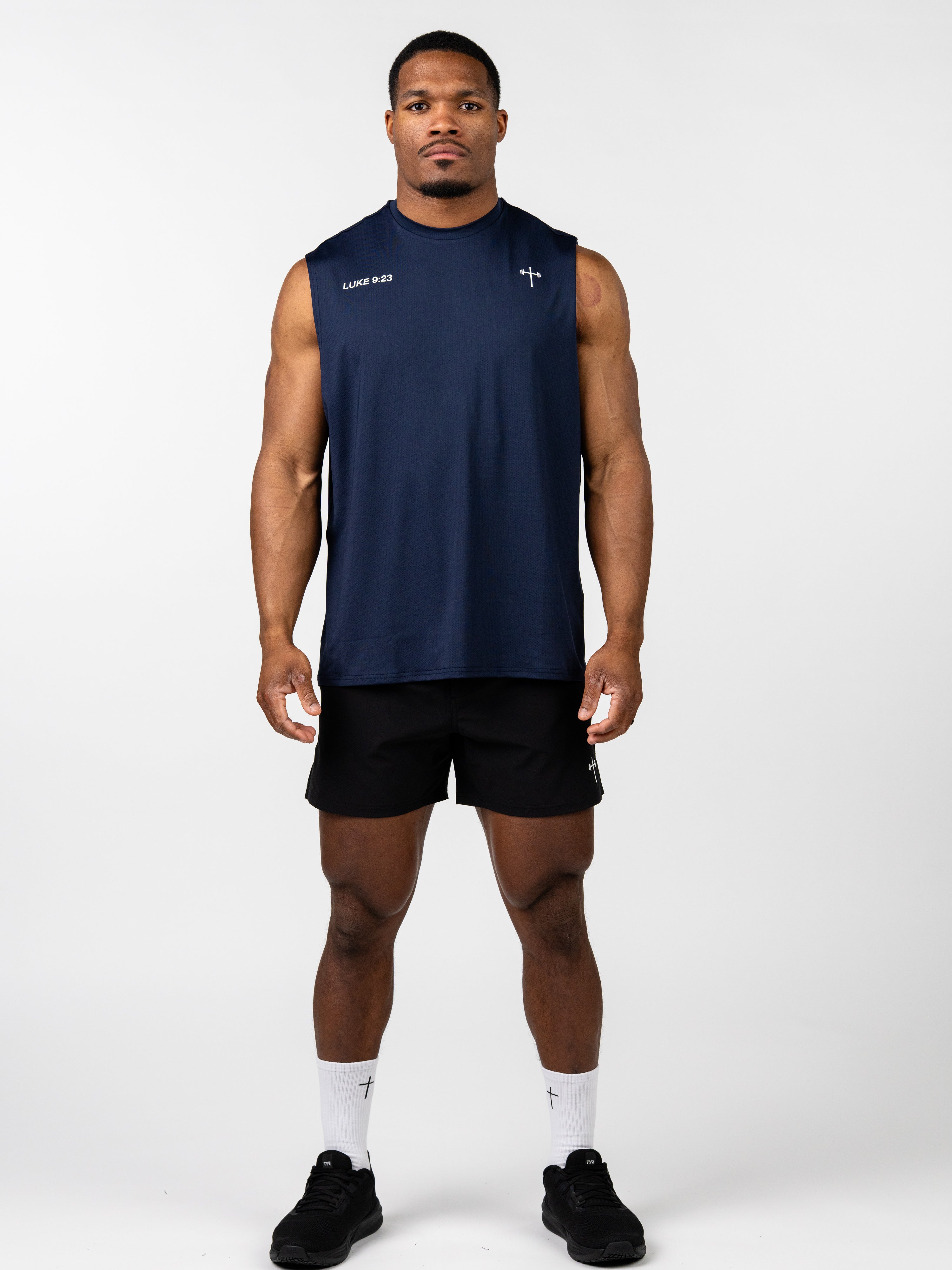 Man wearing a navy blue Carry Your Cross Premium Oversized Tank with subtle cross and scripture detail, paired with black shorts, white socks, and black athletic shoes in a studio setting.