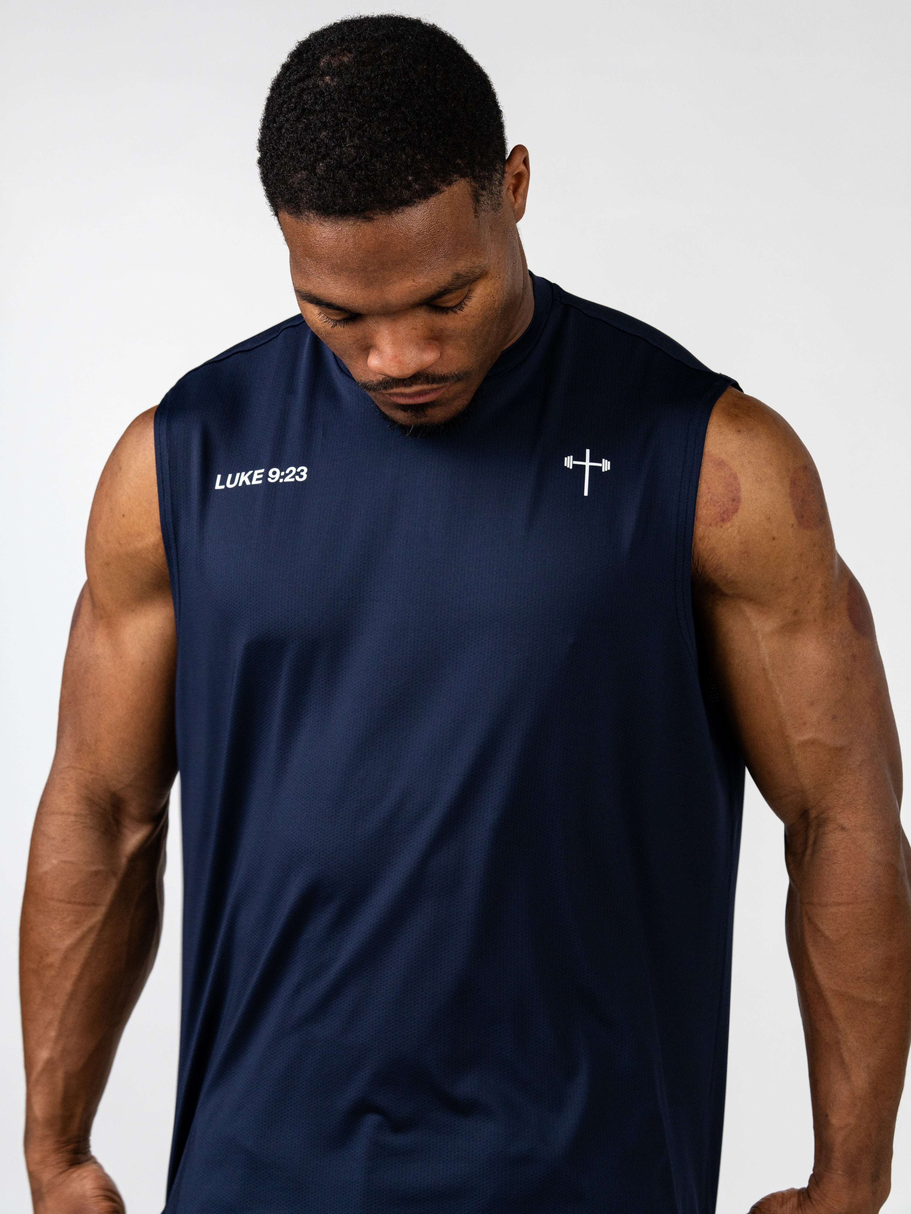 Man wearing a navy blue Carry Your Cross Premium Oversized Tank with a small cross and barbell graphic on the chest and white text reading Luke 9:23, showcasing Christian fitness apparel.
