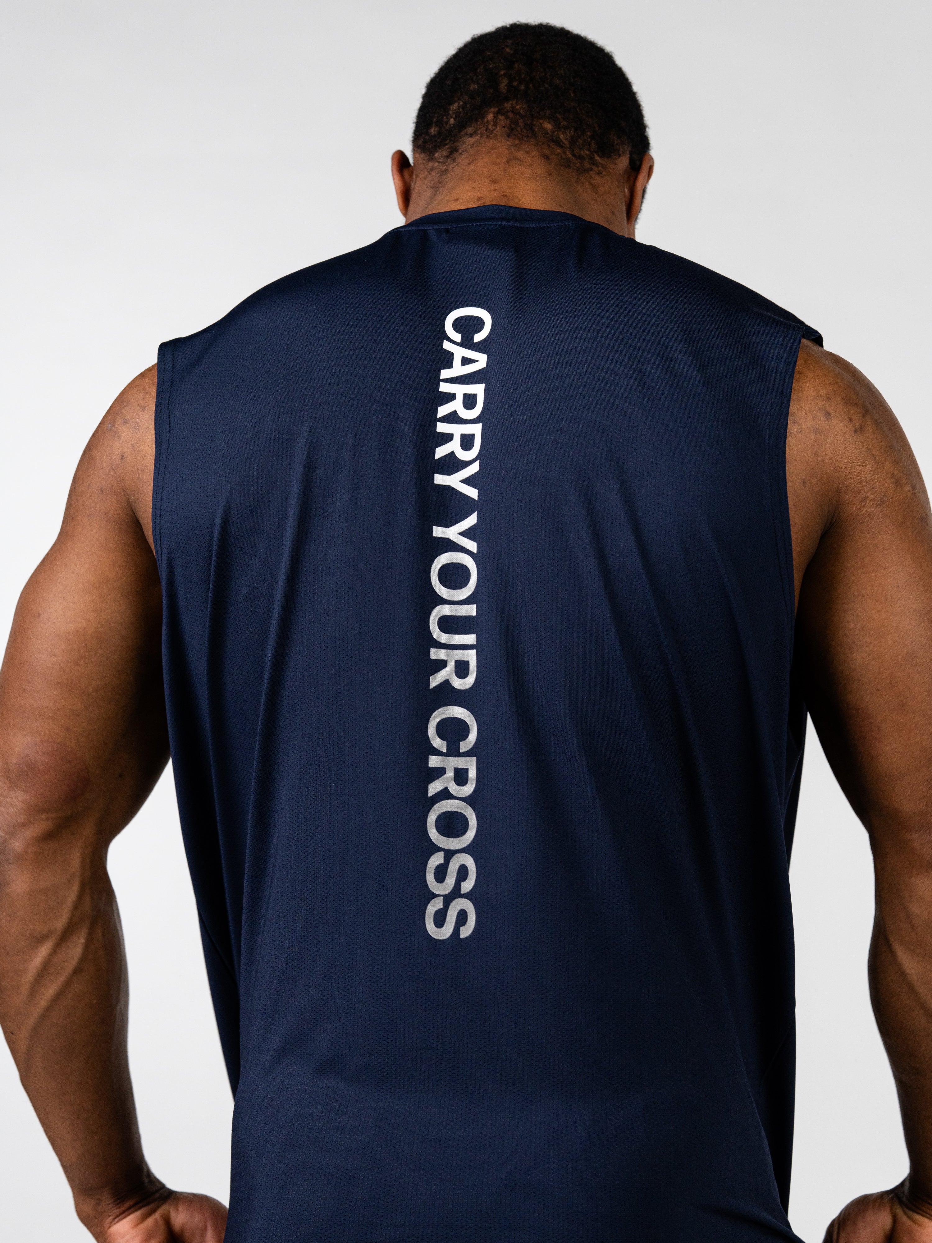 Rear view of a muscular man wearing a navy blue Carry Your Cross Premium Oversized Tank with bold vertical text, highlighting Christian fitness apparel for faith-based workouts.