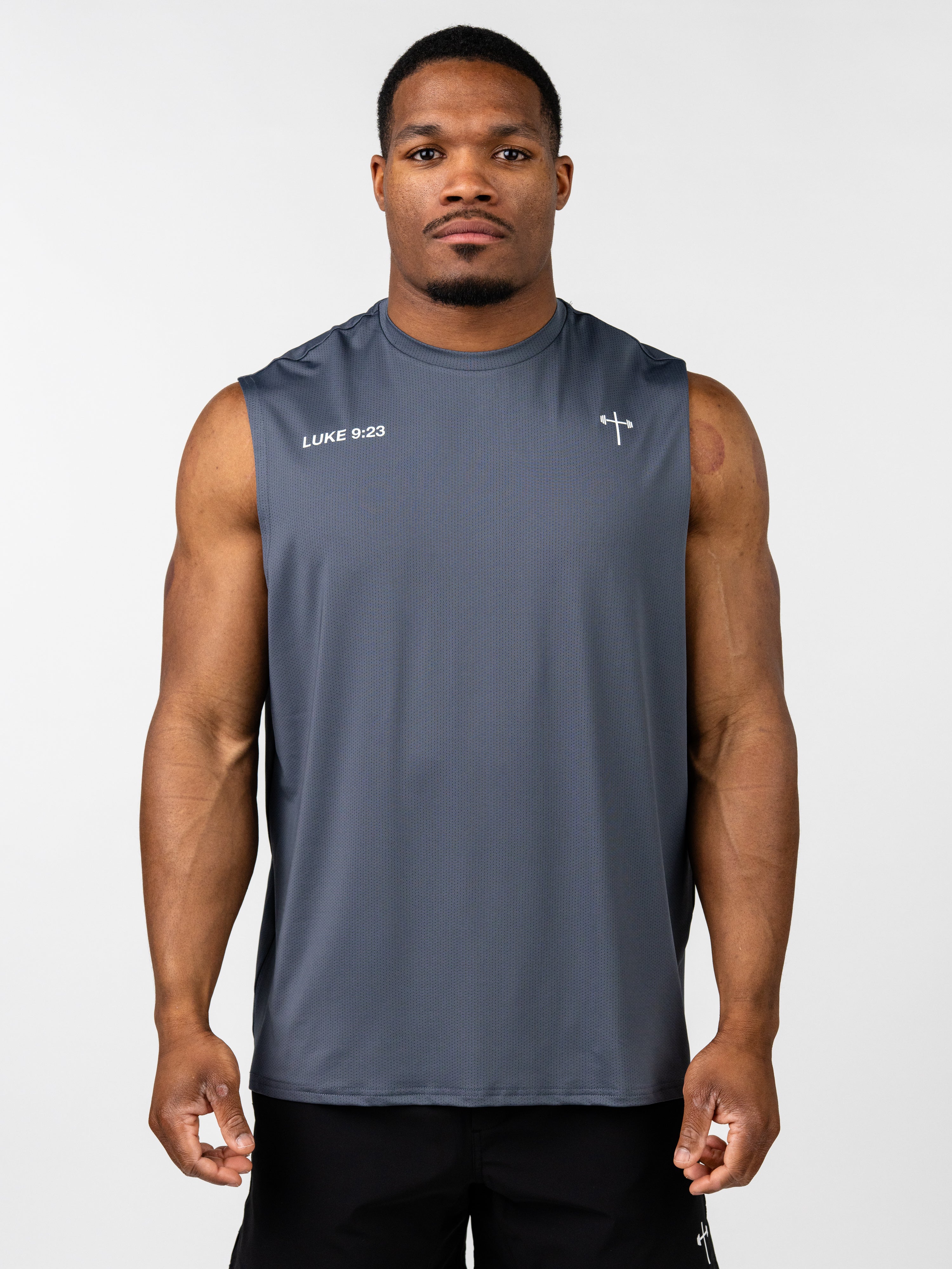 Man wearing a dark gray Carry Your Cross Premium Oversized Tank featuring a small cross with a barbell design and the text "LUKE 9:23" on the upper chest, styled as Christian fitness apparel.