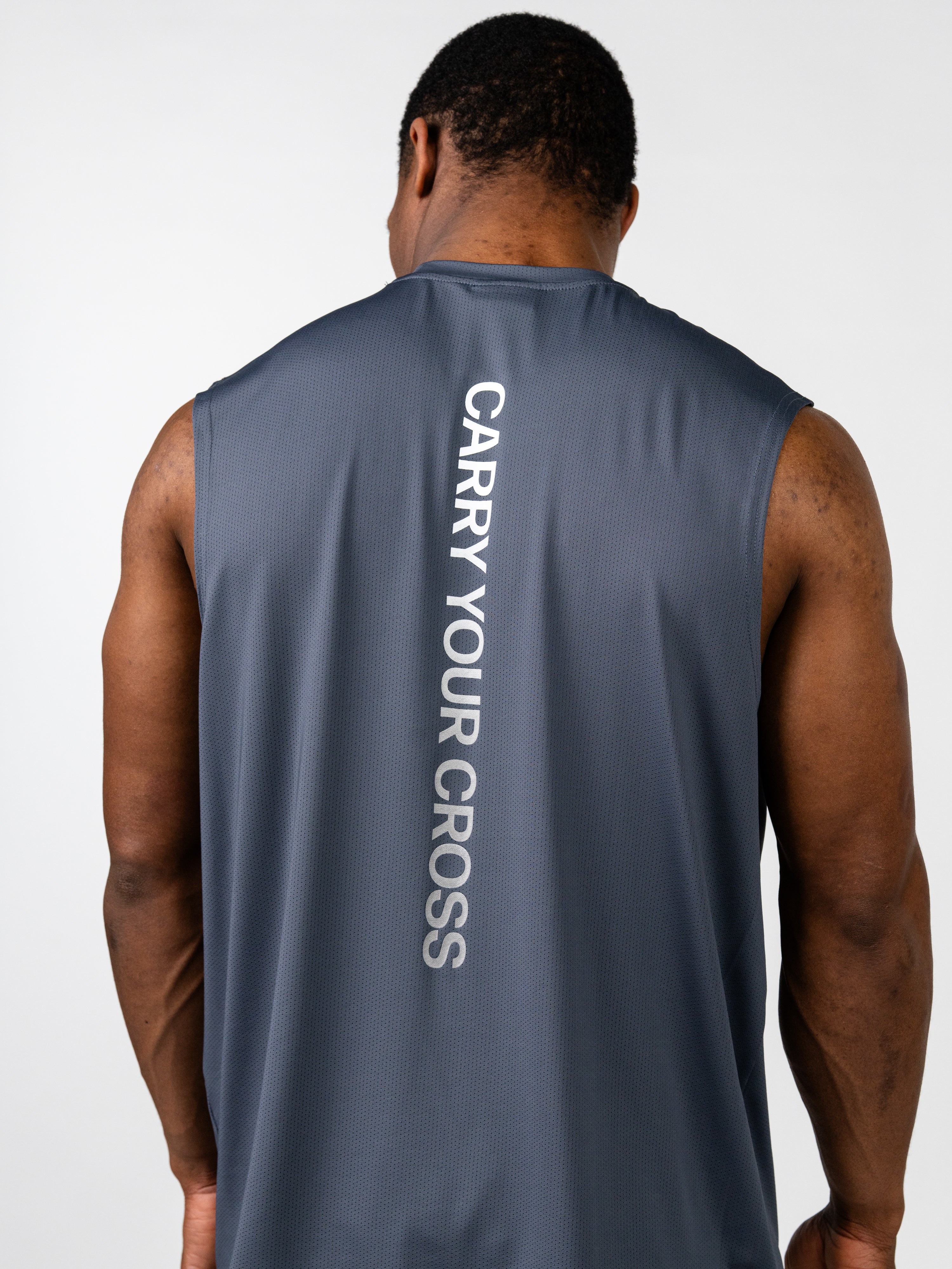 Man wearing the Carry Your Cross Premium Oversized Tank in dark gray, showing the back with "Carry Your Cross" printed vertically in white, highlighting Christian fitness apparel.