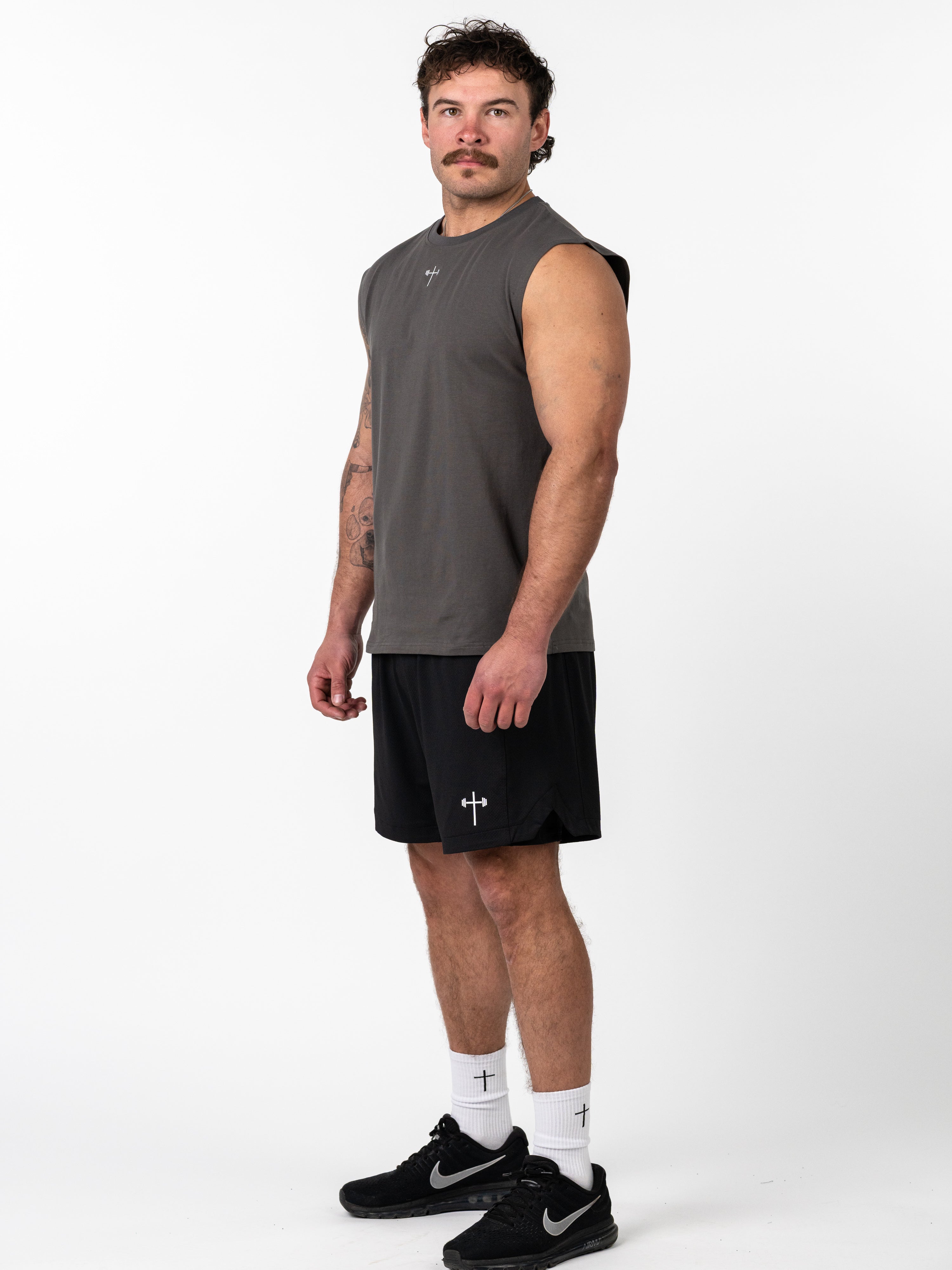 Male athlete wearing Scripture Premium Sleeveless Tank in iron grey, paired with black athletic shorts and white socks featuring a cross and barbell design, standing against a white background.