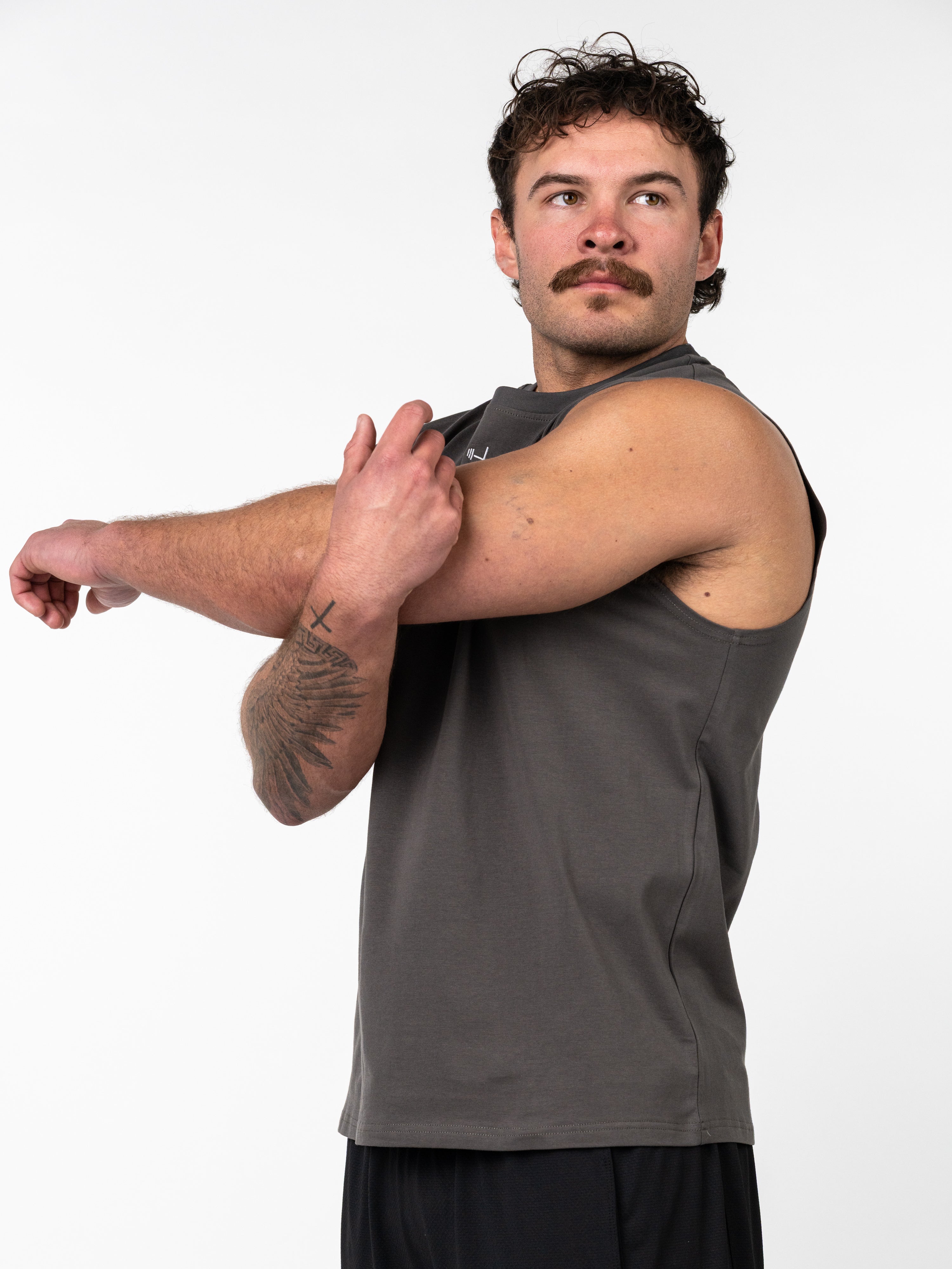 Man wearing a charcoal Scripture Premium Sleeveless Tank stretching his arm, showcasing Christian fitness apparel designed for strength and faith-based training.