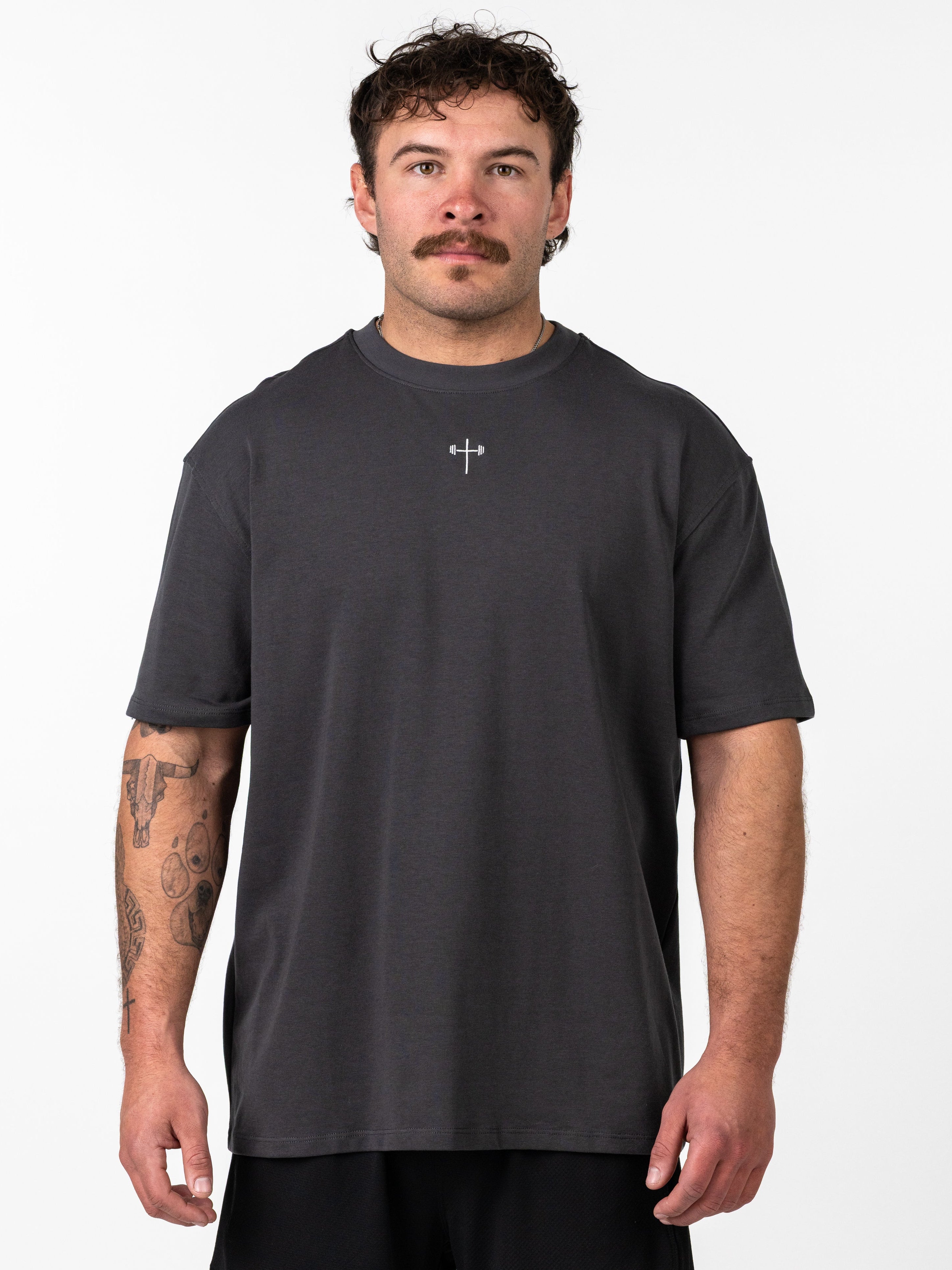 Man wearing a dark gray Classic Oversized Tee featuring a subtle Christian cross and barbell logo on the chest, showcasing a relaxed fit and comfortable activewear style.