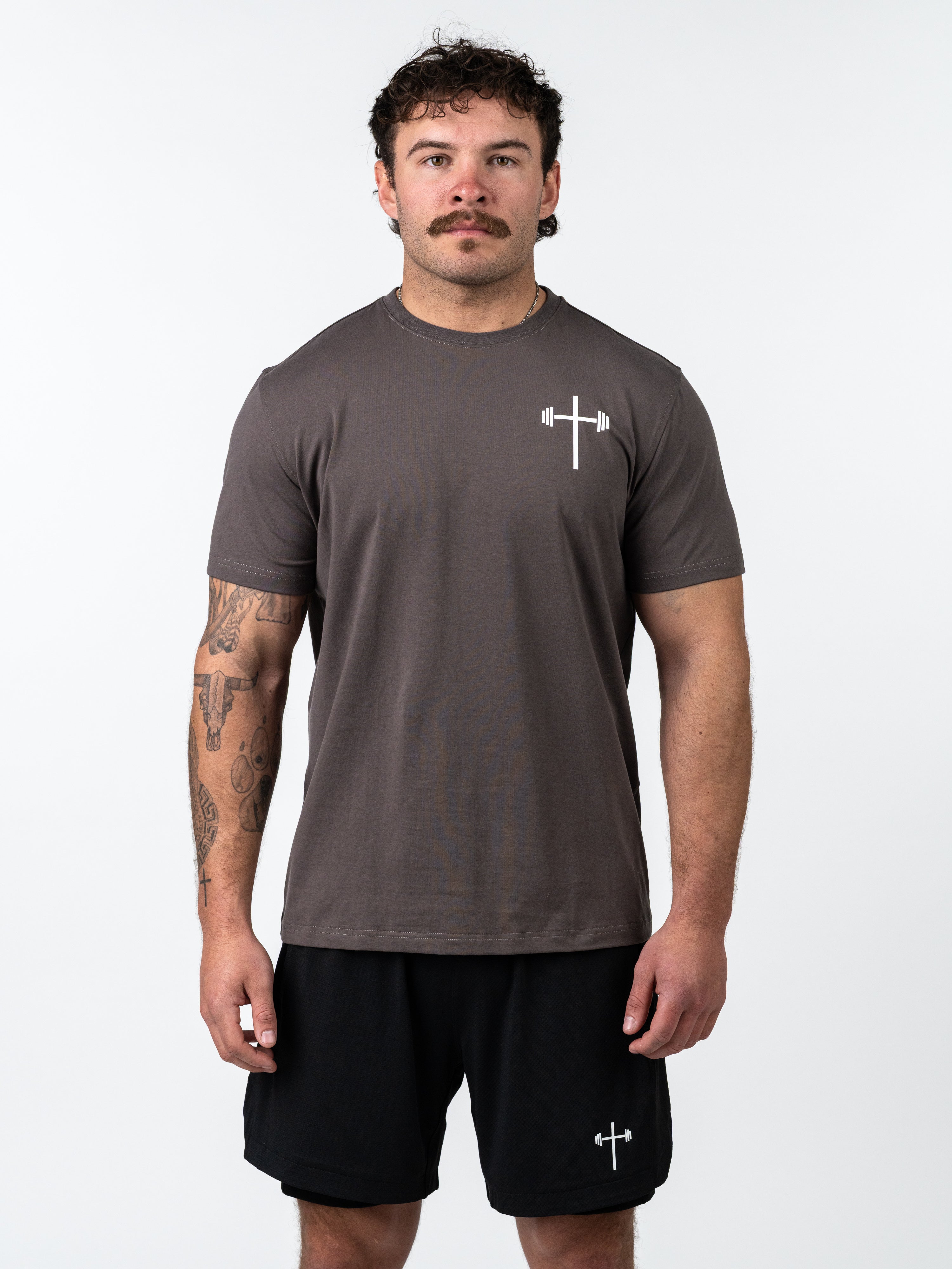 Man wearing a charcoal gray Kingdom Performance Tee featuring a small white cross with barbell logo on the chest, styled with black athletic shorts against a plain white background.