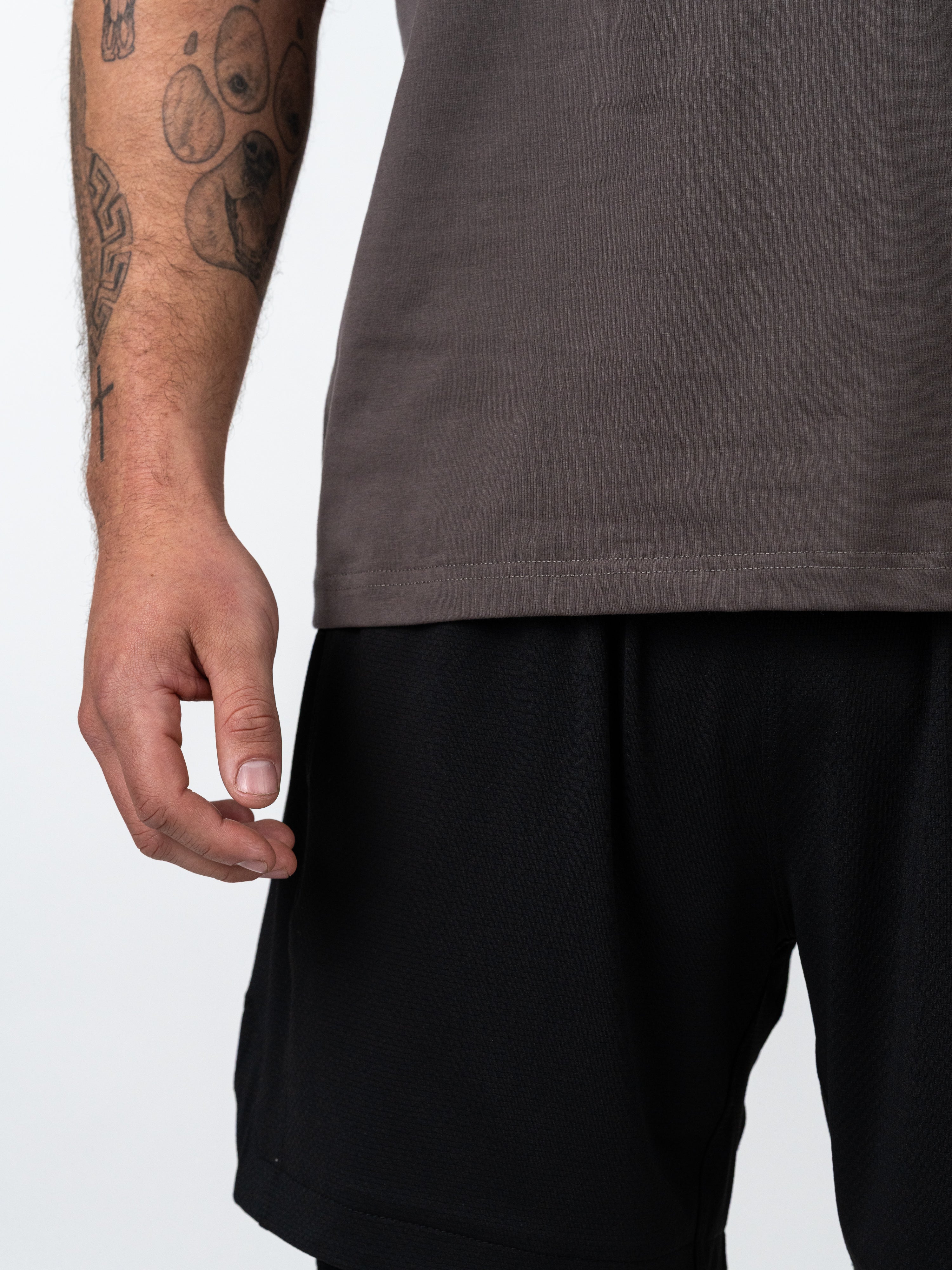Close-up of a person wearing the Kingdom Performance Tee in iron grey, showing the athletic-slim fit fabric and tattoos on the arm, paired with black gym shorts.