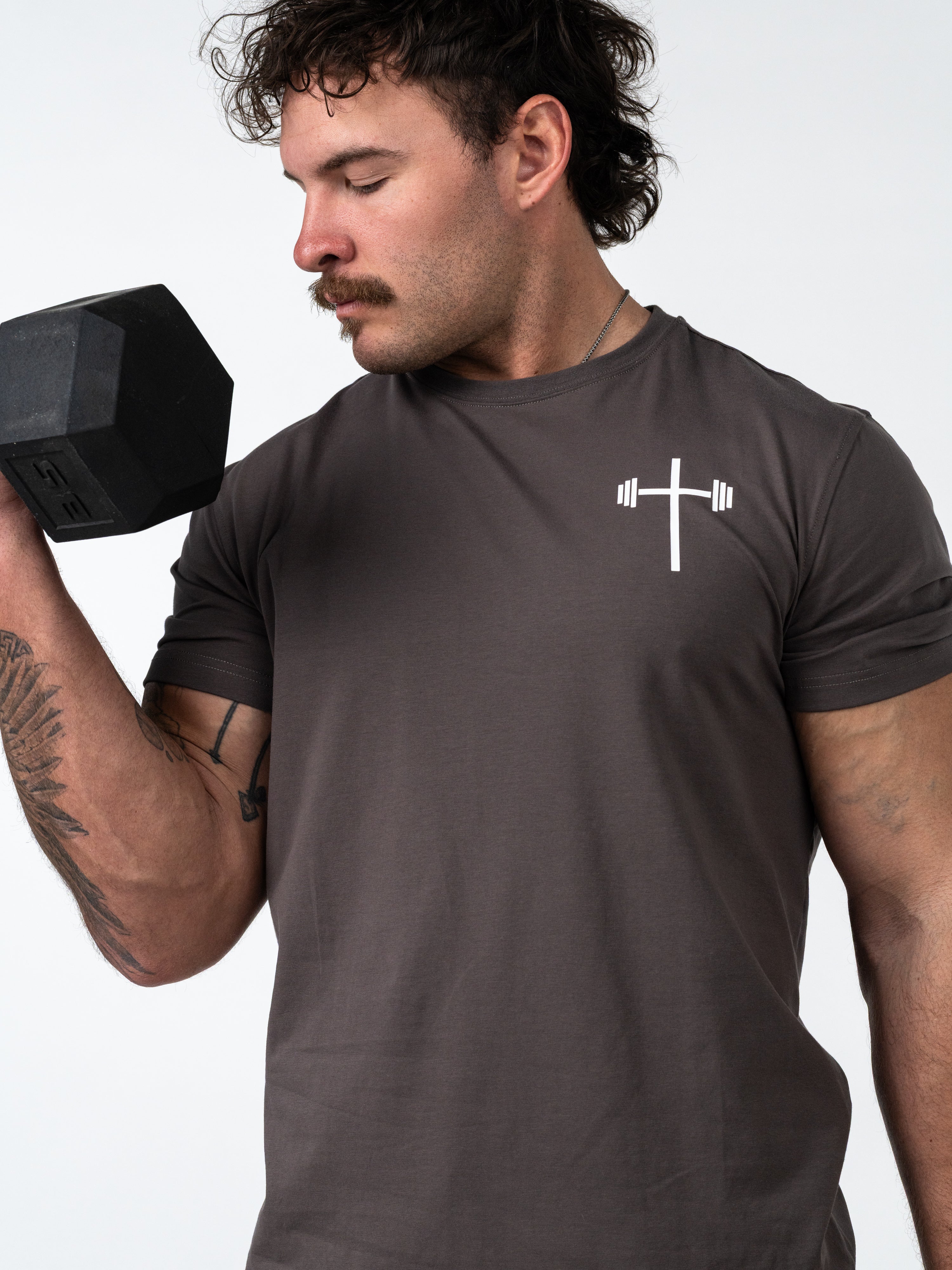 Athletic man lifting dumbbell wearing a dark gray Kingdom Performance Tee with a white cross and barbell design, showcasing Christian fitness apparel for Faith & Fitness enthusiasts.