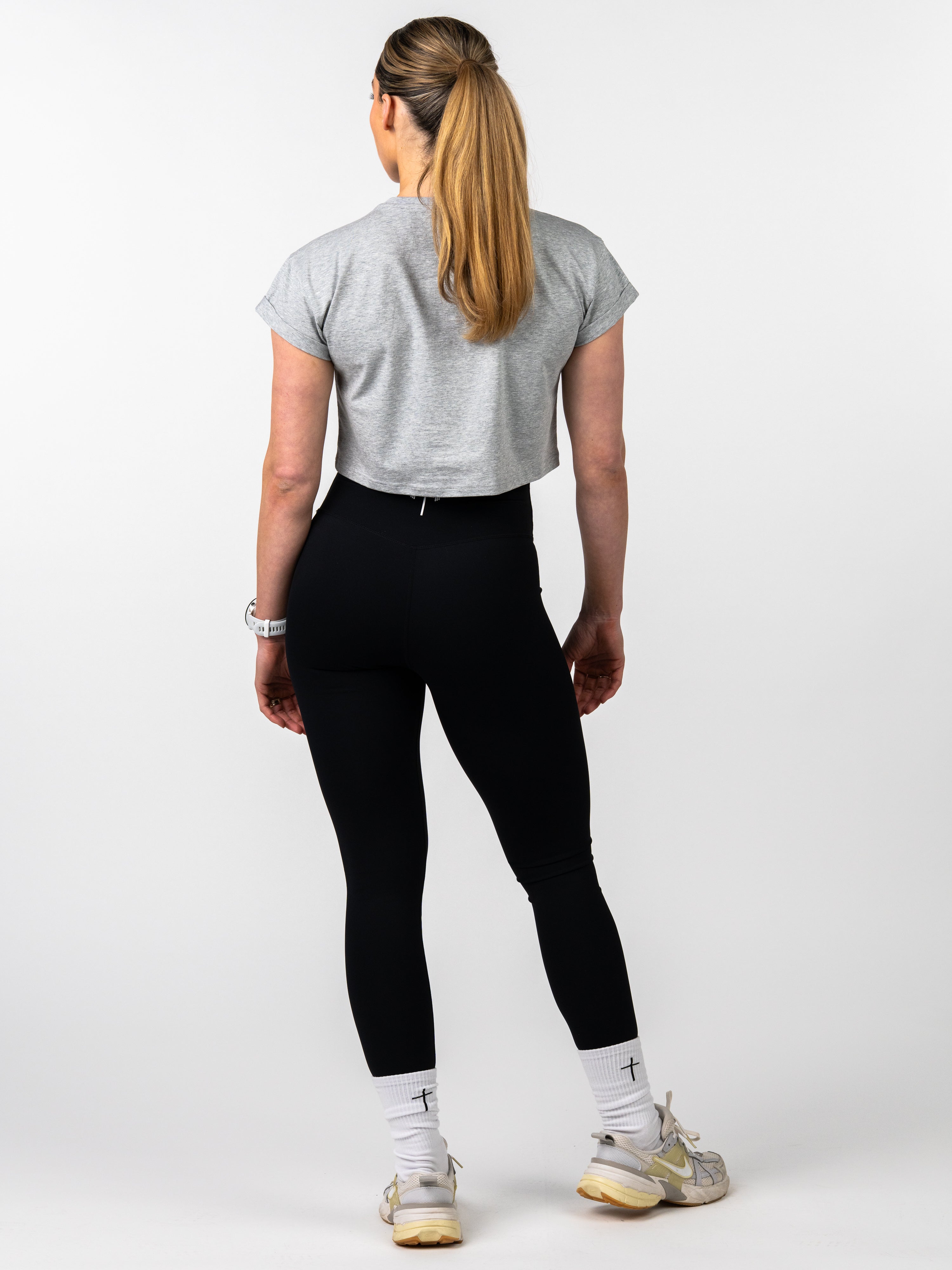 Back view of a woman wearing a gray Jesus is King Essential Crop Tee paired with black leggings, white socks with cross detail, and light-colored athletic shoes, standing against a white background.