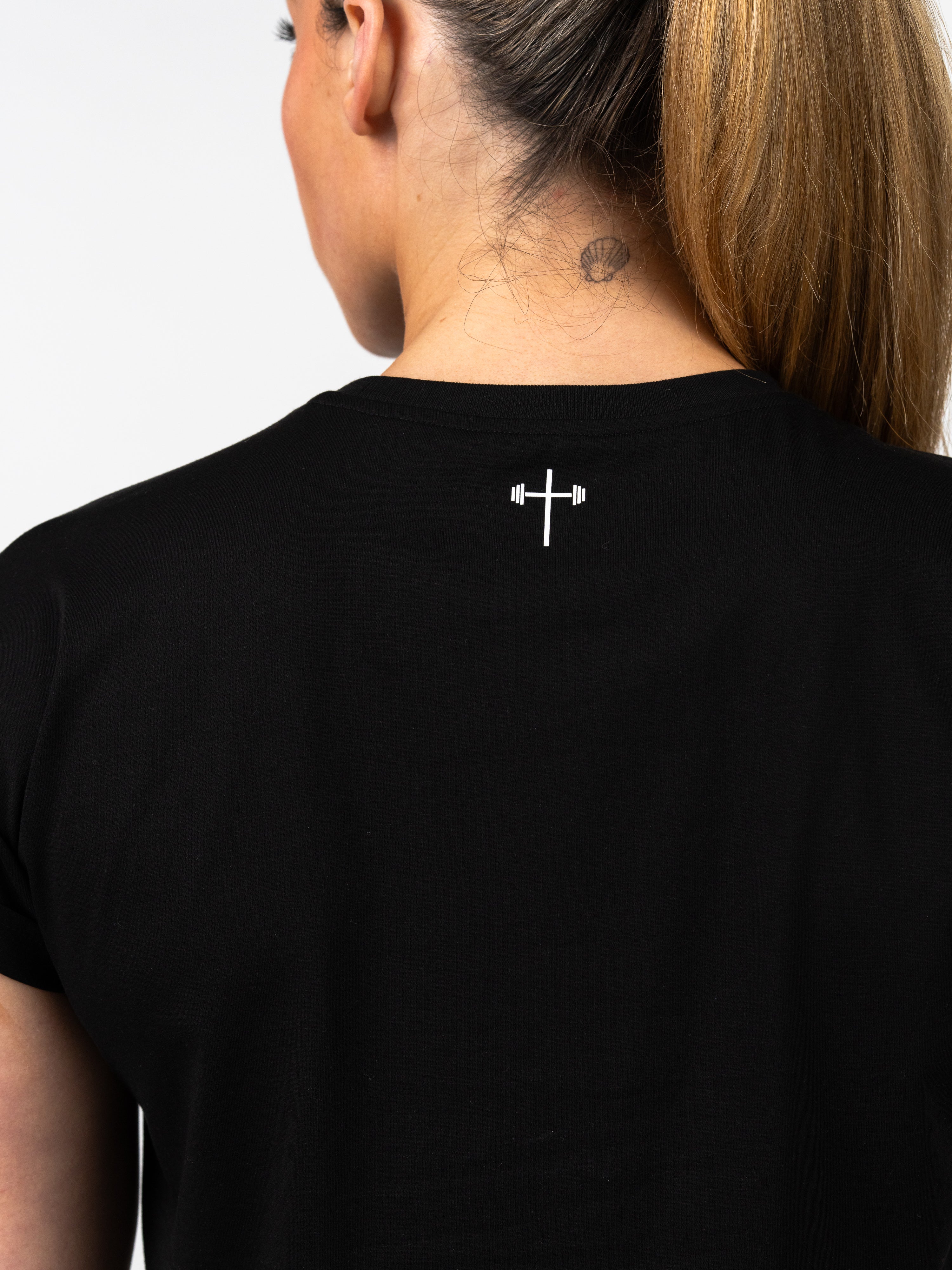 Close-up of a woman wearing a black Jesus is King Essential Crop Tee, showing the back neckline with a small white graphic of a cross incorporated with a barbell, representing Christian fitness apparel.