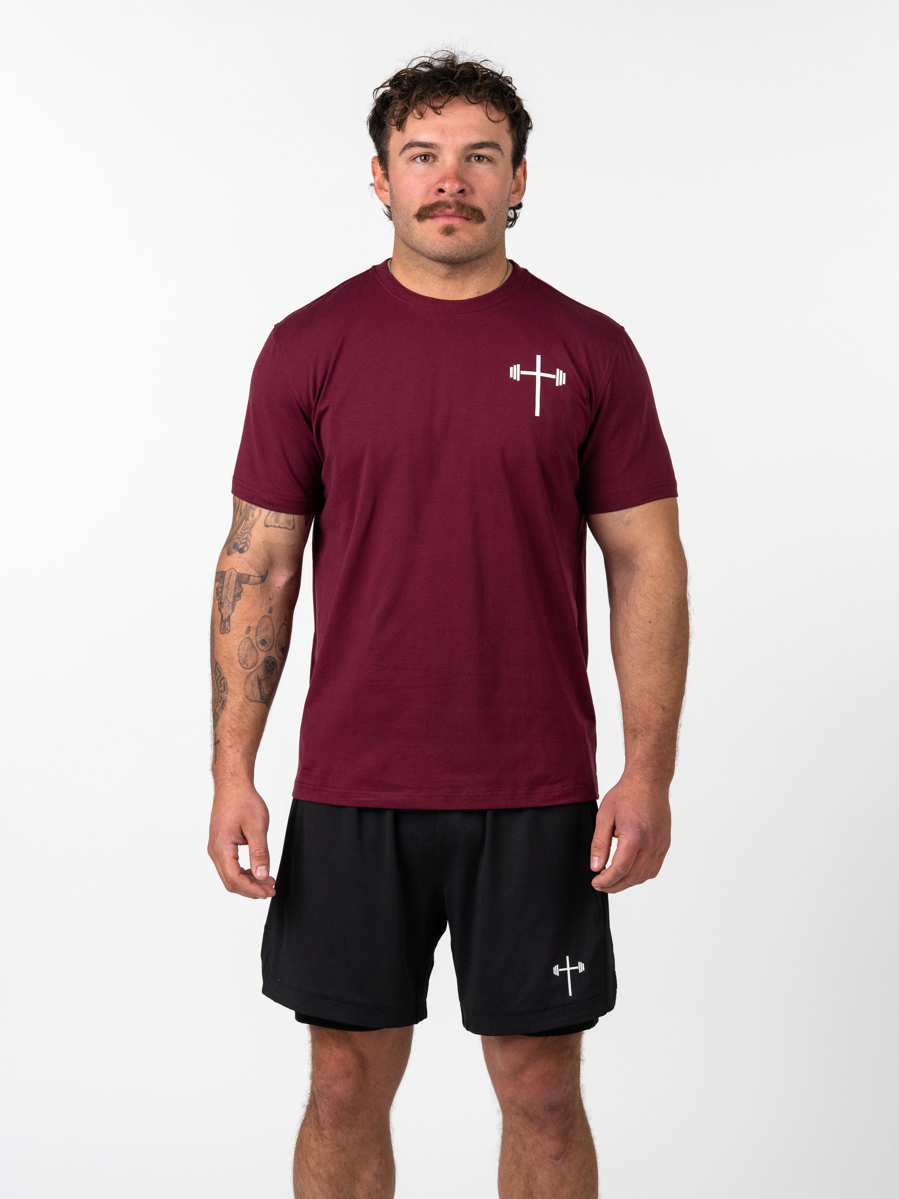 Male model wearing a maroon Kingdom Performance Tee with a white barbell cross logo on the chest, paired with black shorts featuring the same logo, showcasing Christian fitness apparel.