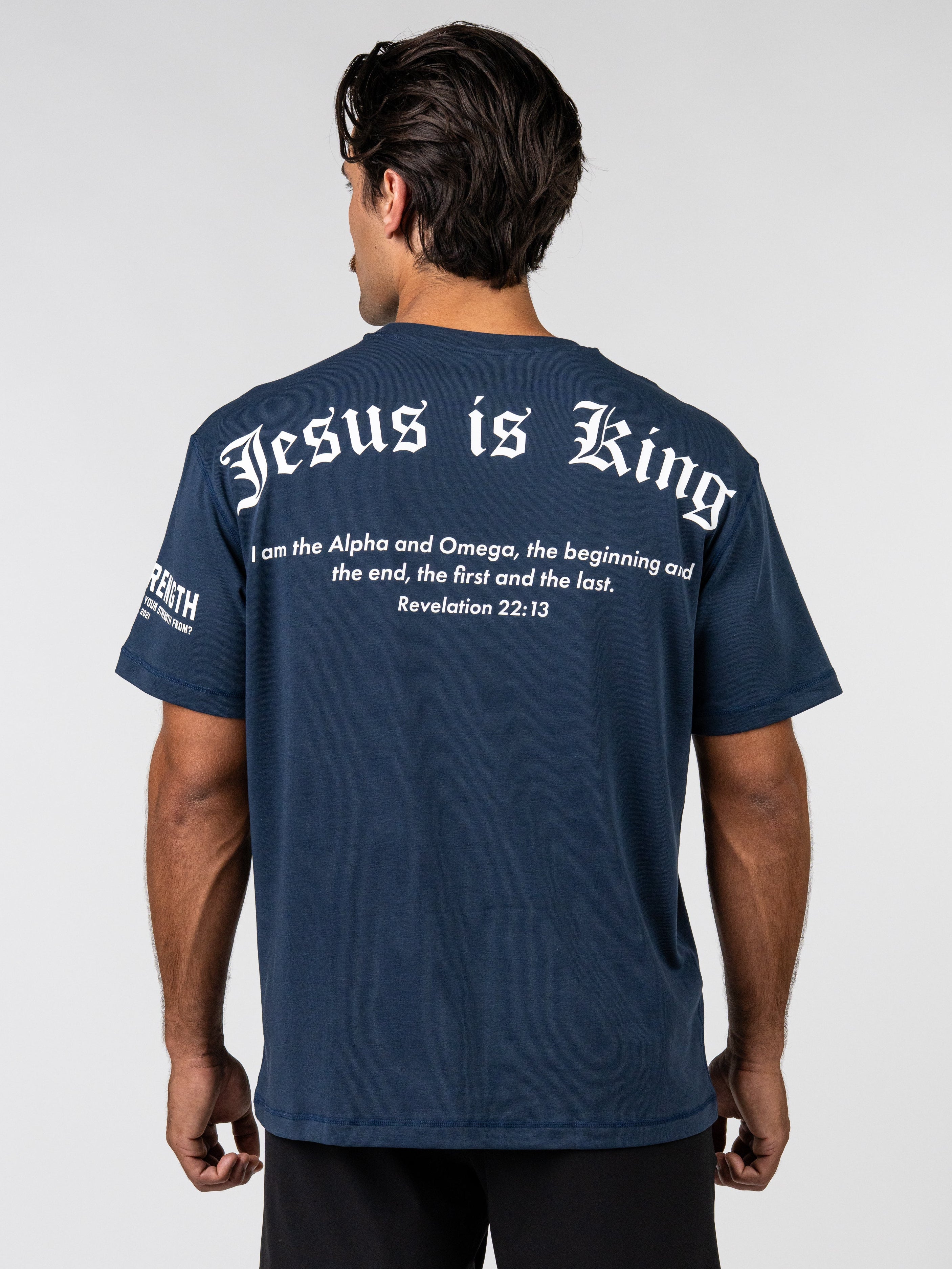 Man wearing a navy Premium Oversized Scripture Tee with white text on the back reading "Jesus is King" and a biblical quote from Revelation 22:13, styled as Christian fitness apparel.