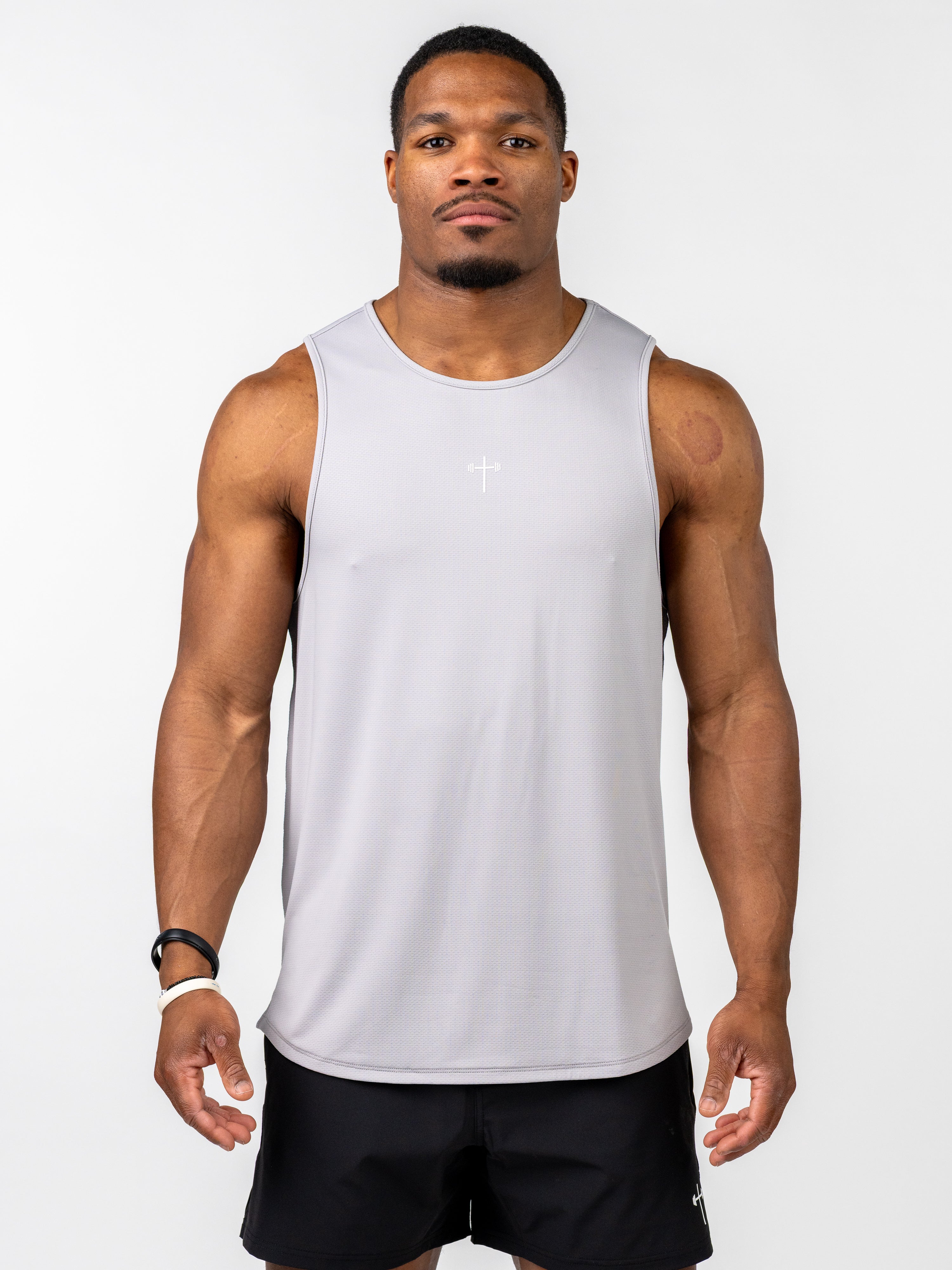 Athletic man wearing a light grey Signature Tank with a small white cross and barbell design on the chest, paired with black shorts, standing against a plain white background.