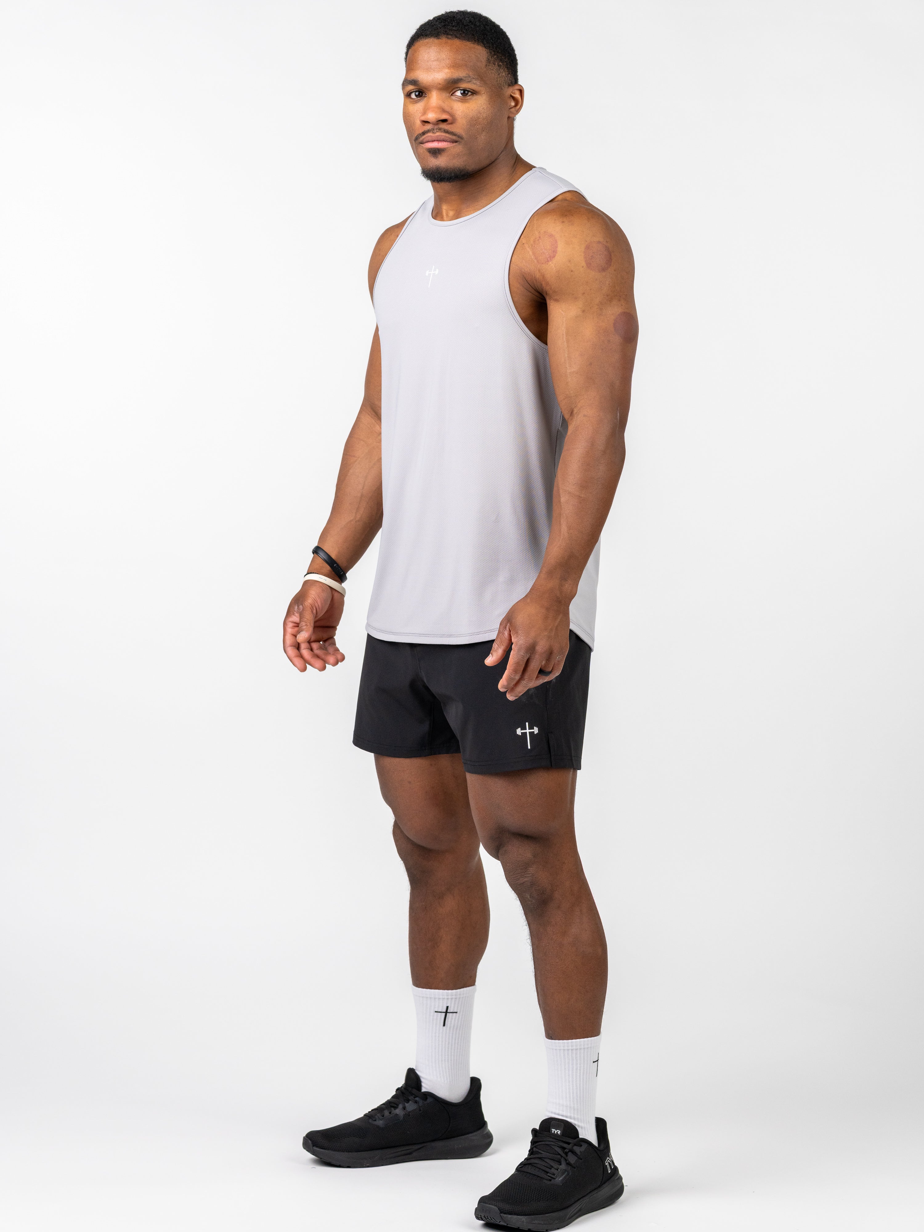 Man wearing a light grey Signature Tank with a small cross and barbell design, paired with black shorts, white socks featuring a cross, and black athletic shoes.
