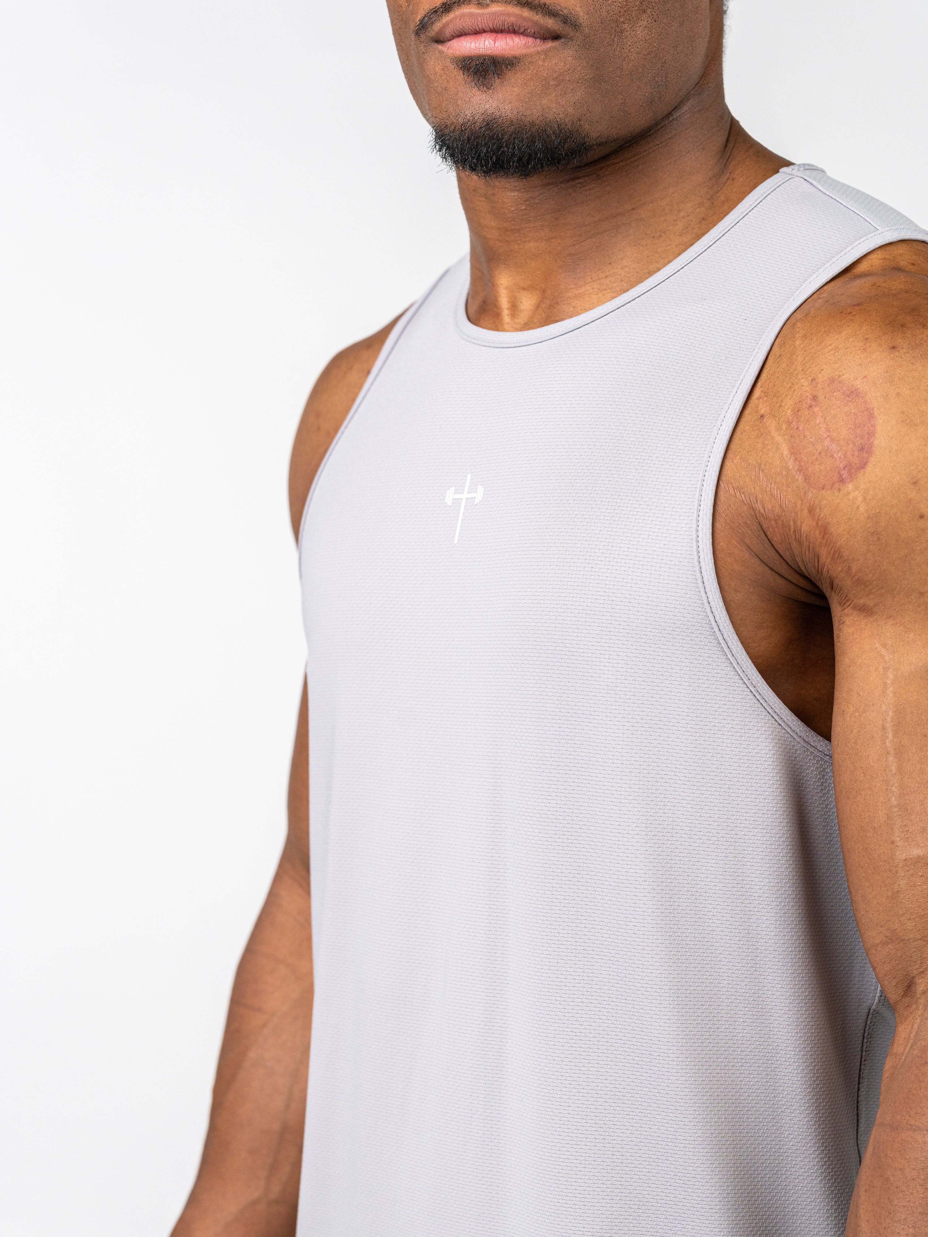 Close-up of a man wearing a light grey Signature Tank made from breathable, stretch fabric, featuring a subtle Christian cross with a barbell design on the chest.