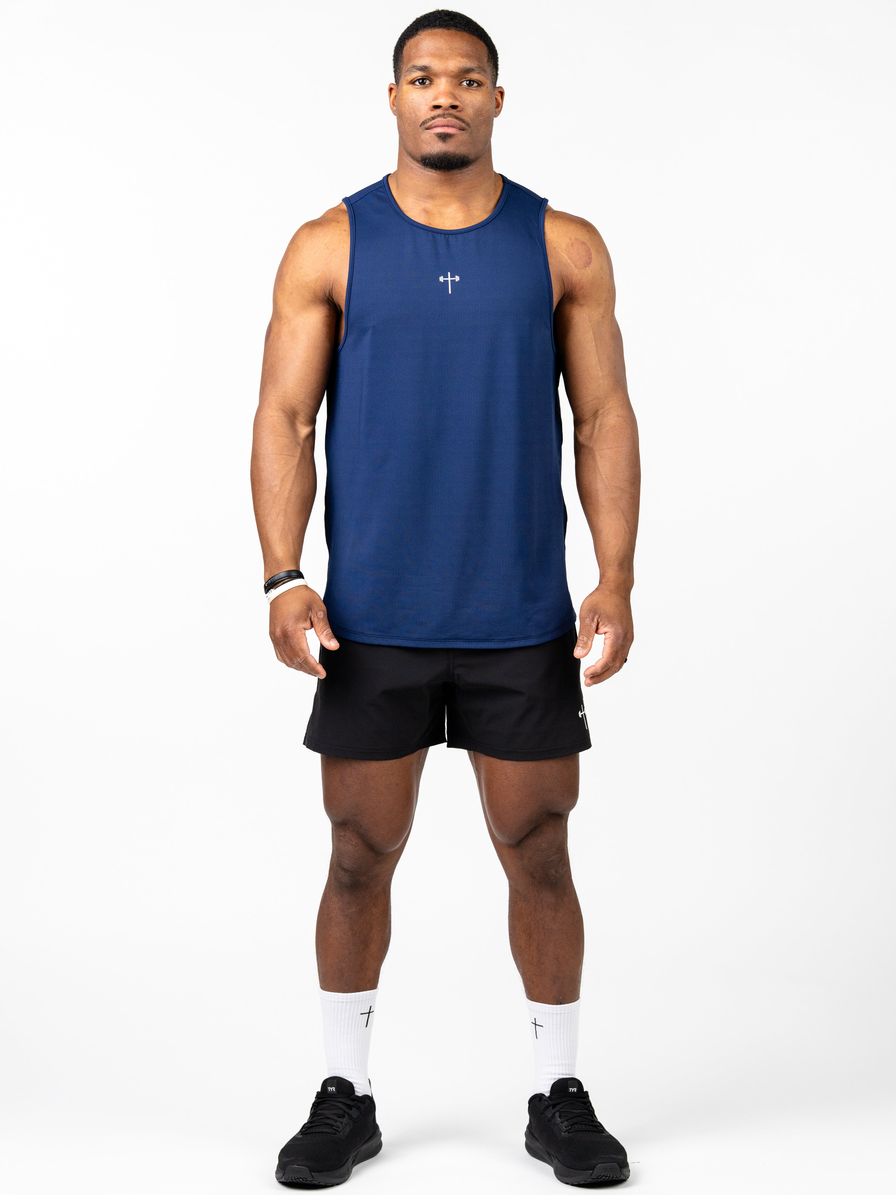 Athletic man wearing a navy blue Signature Tank with a small cross and barbell graphic on the chest, paired with black shorts and white socks, standing against a white background.