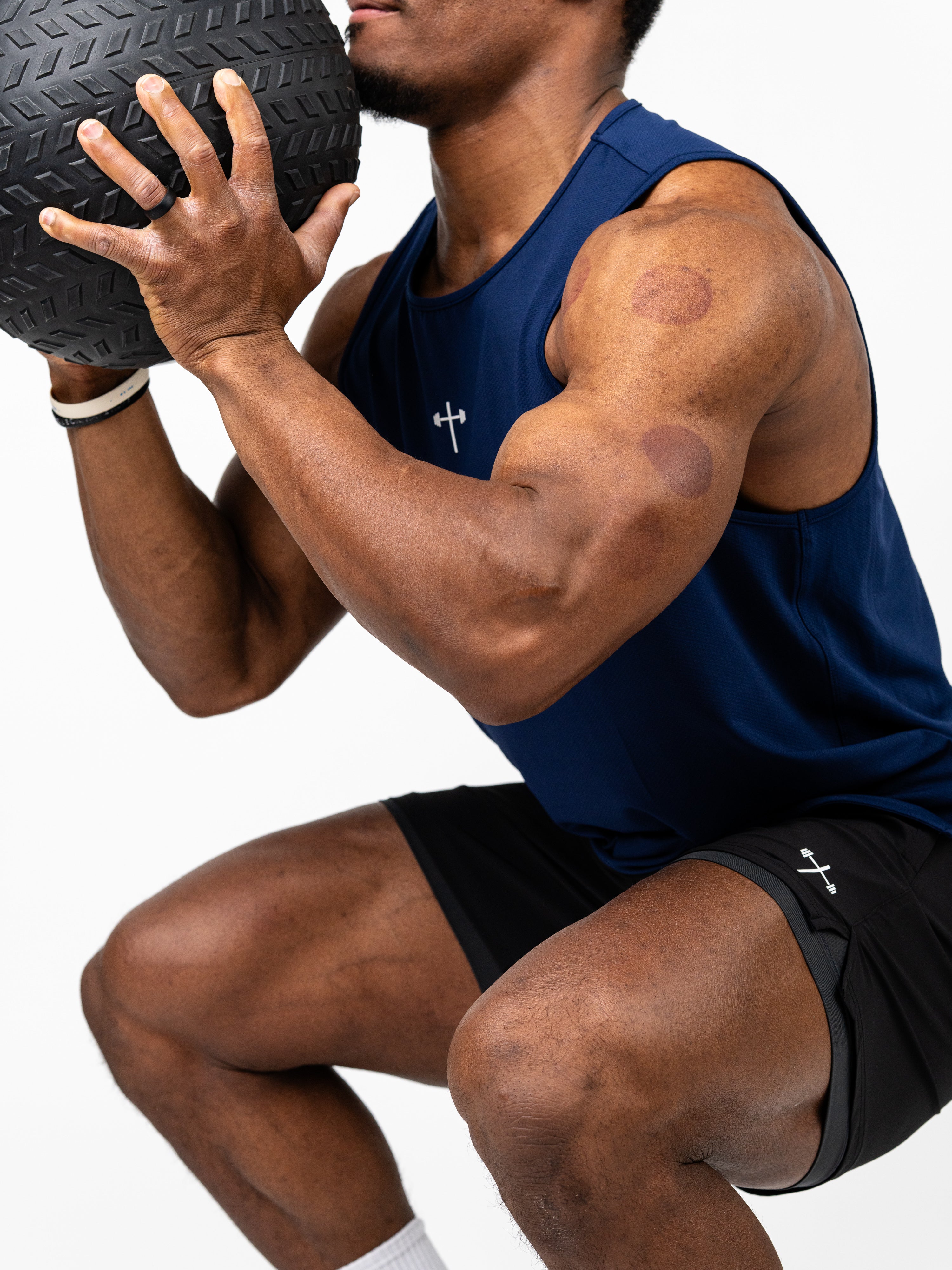 Athletic man squatting while holding a textured medicine ball, wearing a navy blue Signature Tank with a subtle cross and barbell logo, paired with black athletic shorts.