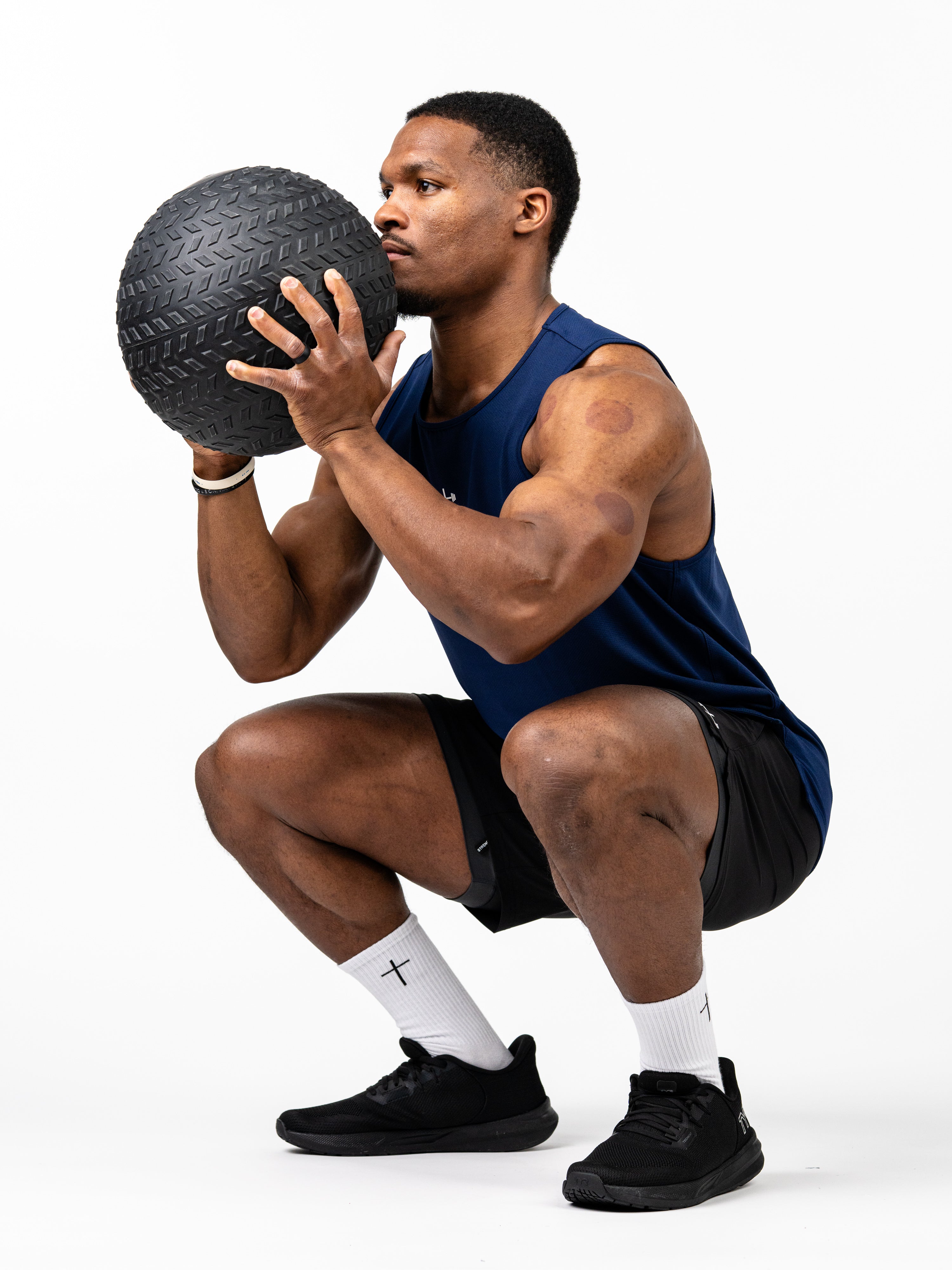 Athletic man wearing a navy blue Signature Tank and black shorts performing a squat while holding a textured black medicine ball, showcasing Christian fitness apparel.